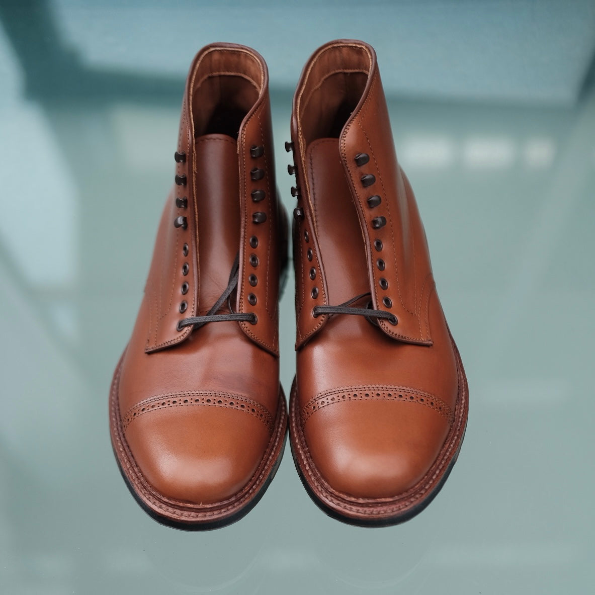 The Alden Madison D2825HC - Jumper Boot in Burnished Tan Calfskin is showcased on a reflective glass surface, featuring black laces, classic design with toe cap detailing, and a sturdy commando sole.