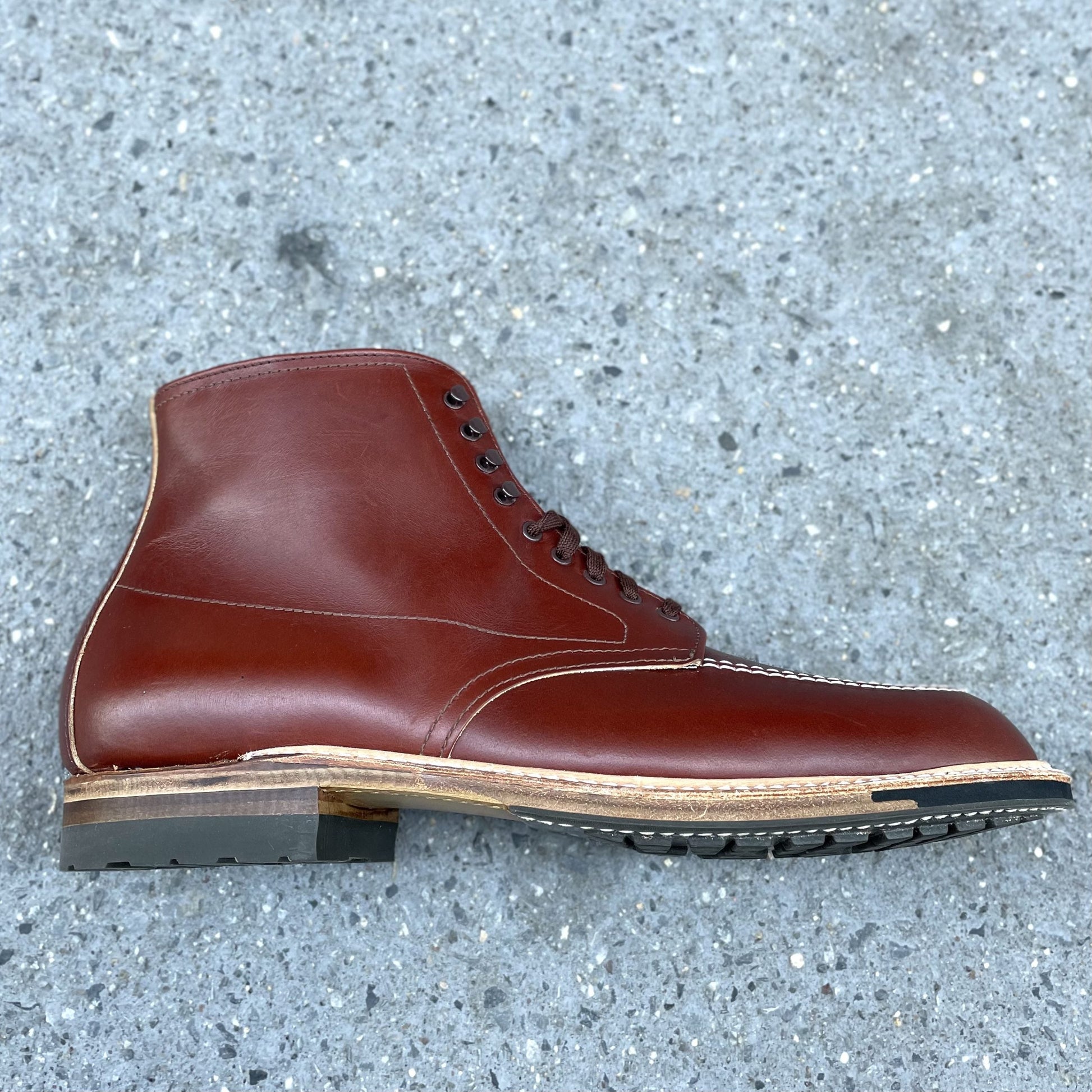 A single Alden Madison 405C - Original 405 Indy Brown leather boot with black laces rests on gray speckled pavement. It showcases a thick Commando Sole with a wood-like accent layer and visible stitching along the edge.