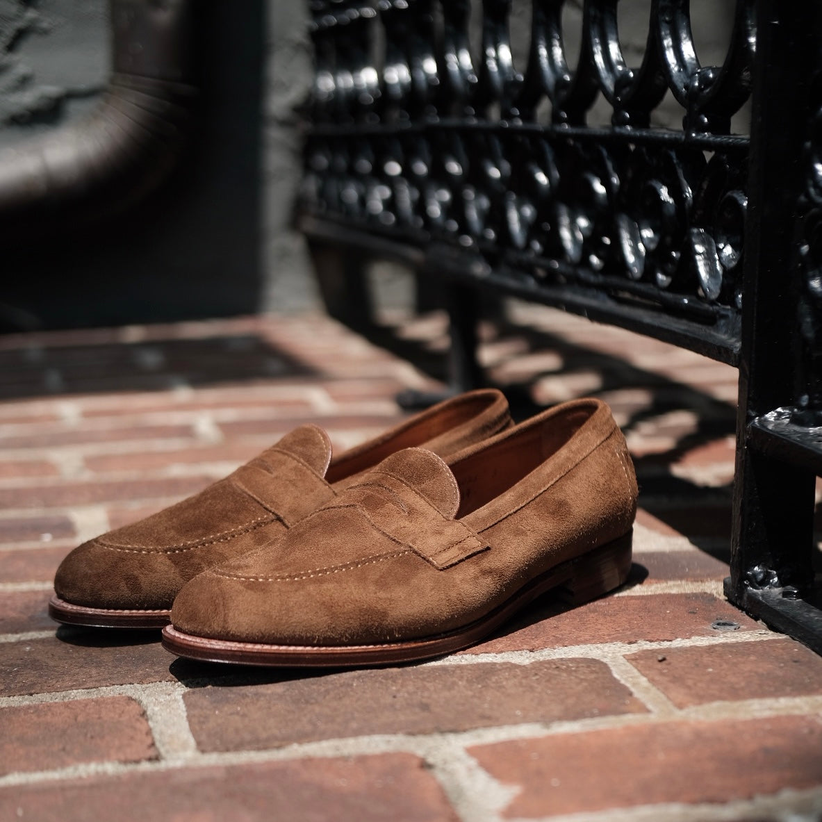 The Alden Madison 96941F Flex Penny Loafer in Snuff Suede, crafted on the Copley Last, rests elegantly on a sunlit brick pavement near an ornate black metal railing, casting delicate shadows.