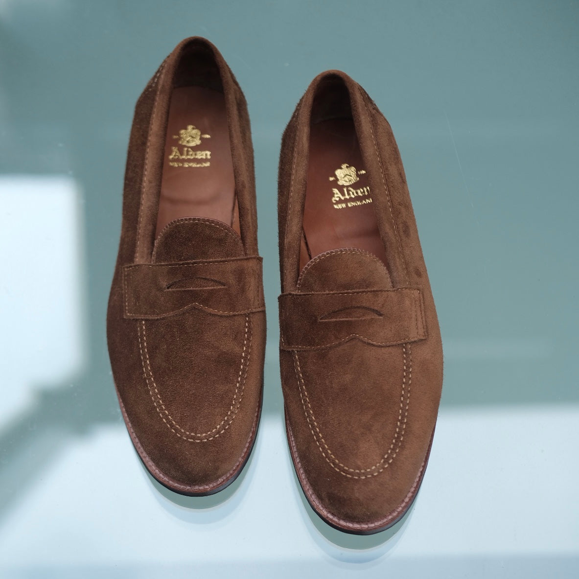 The 96941F - Flex Penny Loafer by Alden Madison features a subtle bow and is crafted from rich Snuff Suede. Displayed on a reflective surface, each shoe has the brand name Alden printed in gold inside.