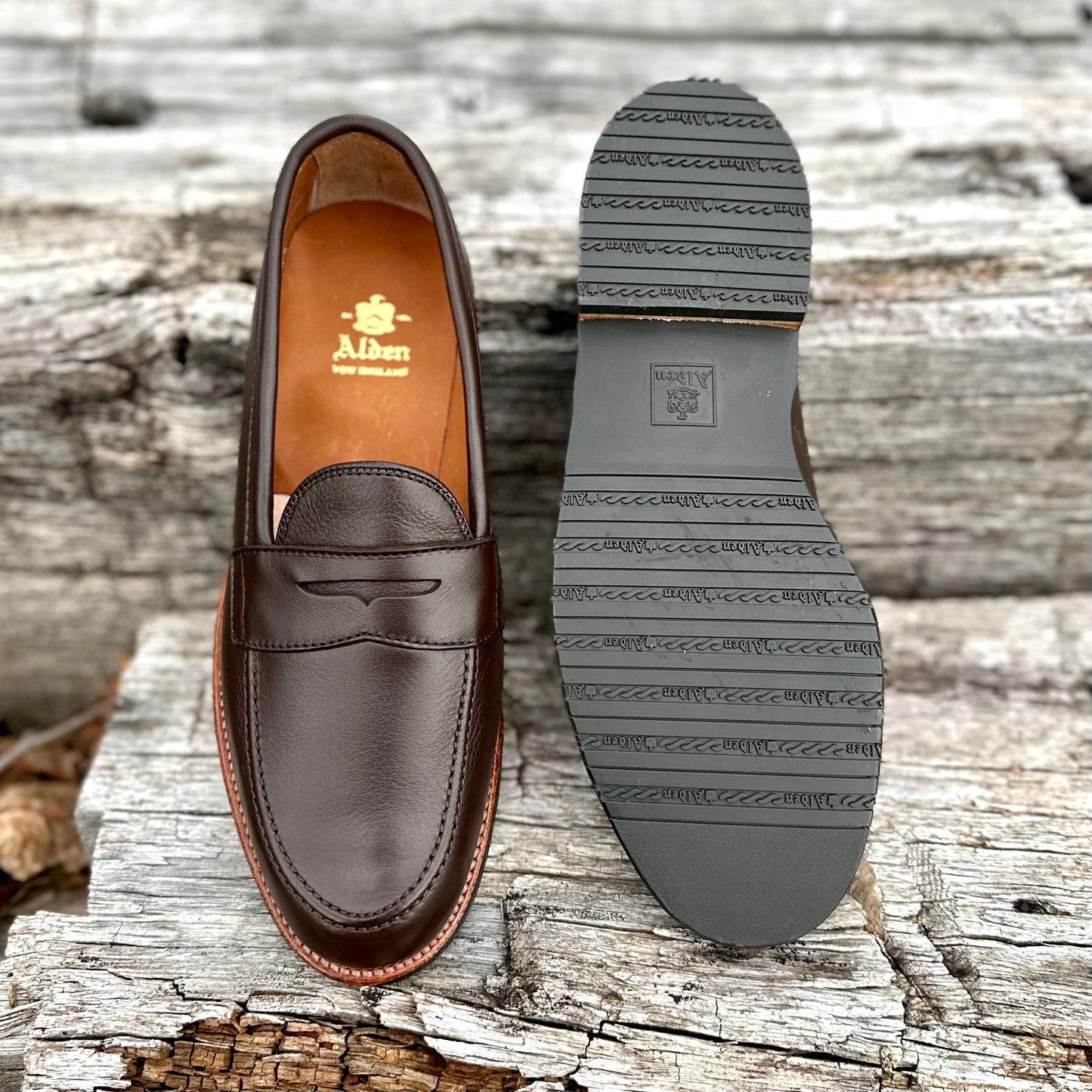 The Alden Madison D2203L - Penny Loafer in Brown Lady Calf features a penny slot, a gold logo on the insole, Antique Flat Welt, and textured black rubber sole for grip, all set on weathered wooden planks.
