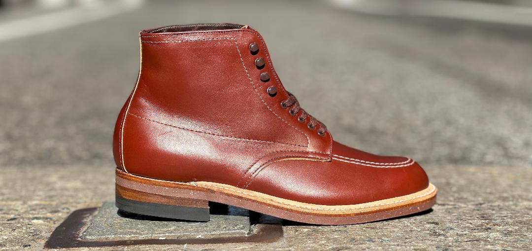 A single Alden Madison 405 - Indy Boot (Original) in Mid Brown Calfskin, with a lace-up design, contrasting light brown sole, and dark heel, rests on urban pavement in sunlight. The background is slightly blurred, highlighting the boots craftsmanship and texture.