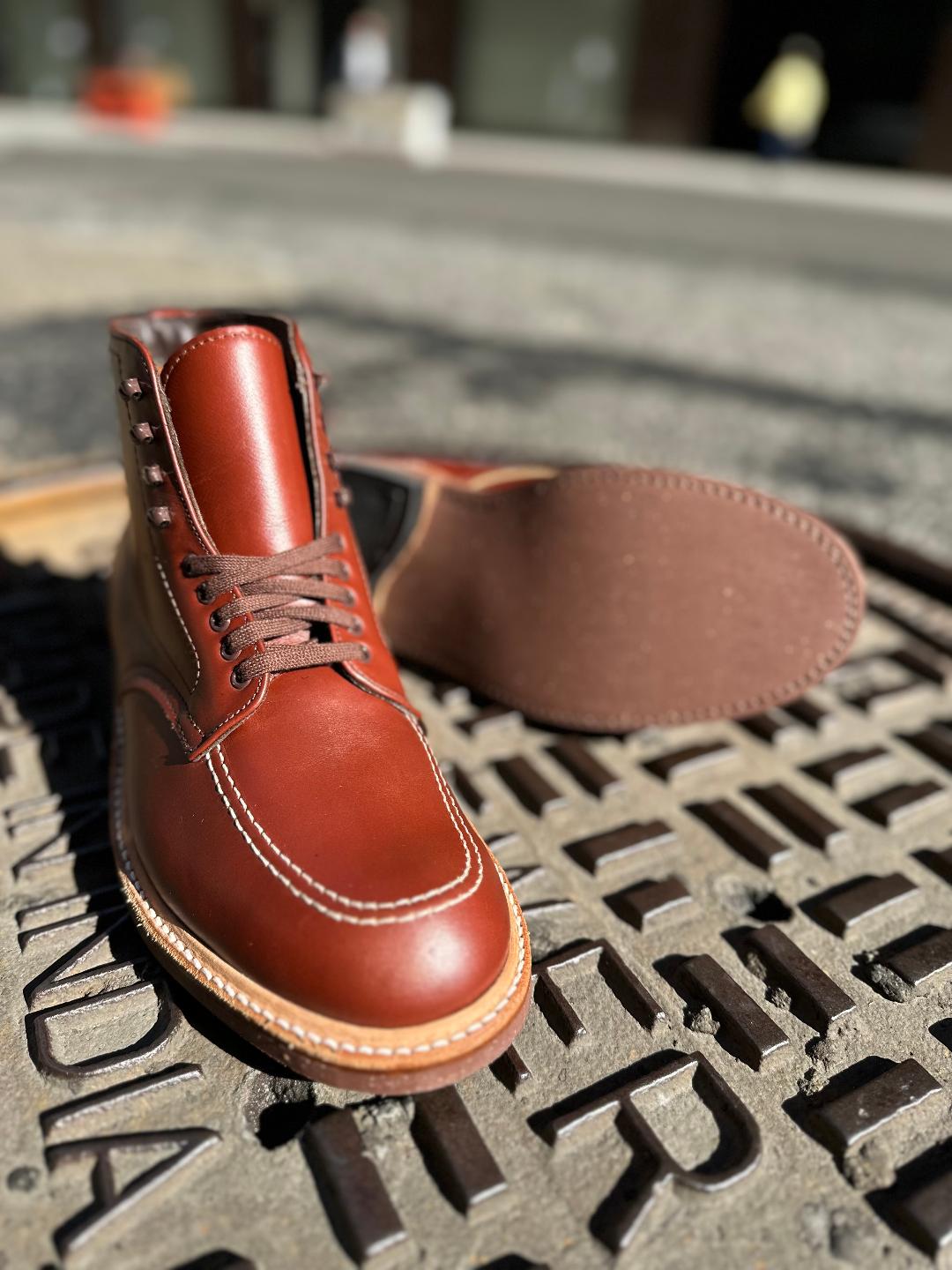 A close-up of Alden Madisons 405 - Indy Boot (Original) in Mid Brown Calfskin shows the upright left boot with visible laces and stitching, while the right boot lies on its side, displaying the sole against a blurred background.
