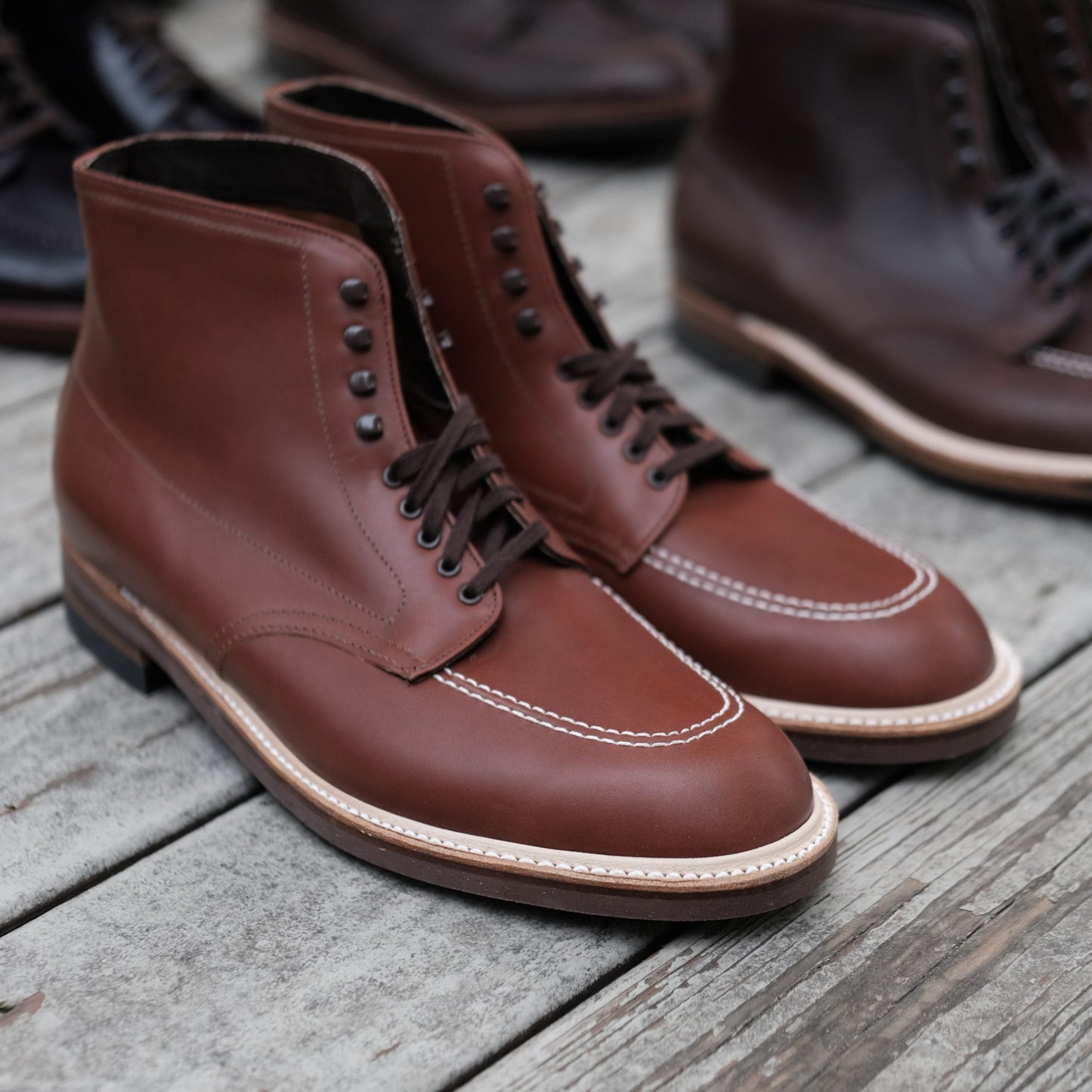 A pair of Alden Madisons 405 - Indy Boots in Mid Brown Calfskin, elegantly crafted with dark laces and white stitching along the toe cap and sole, is showcased on a wooden surface, with more pairs artistically blurred in the background to highlight their rugged yet refined appeal.