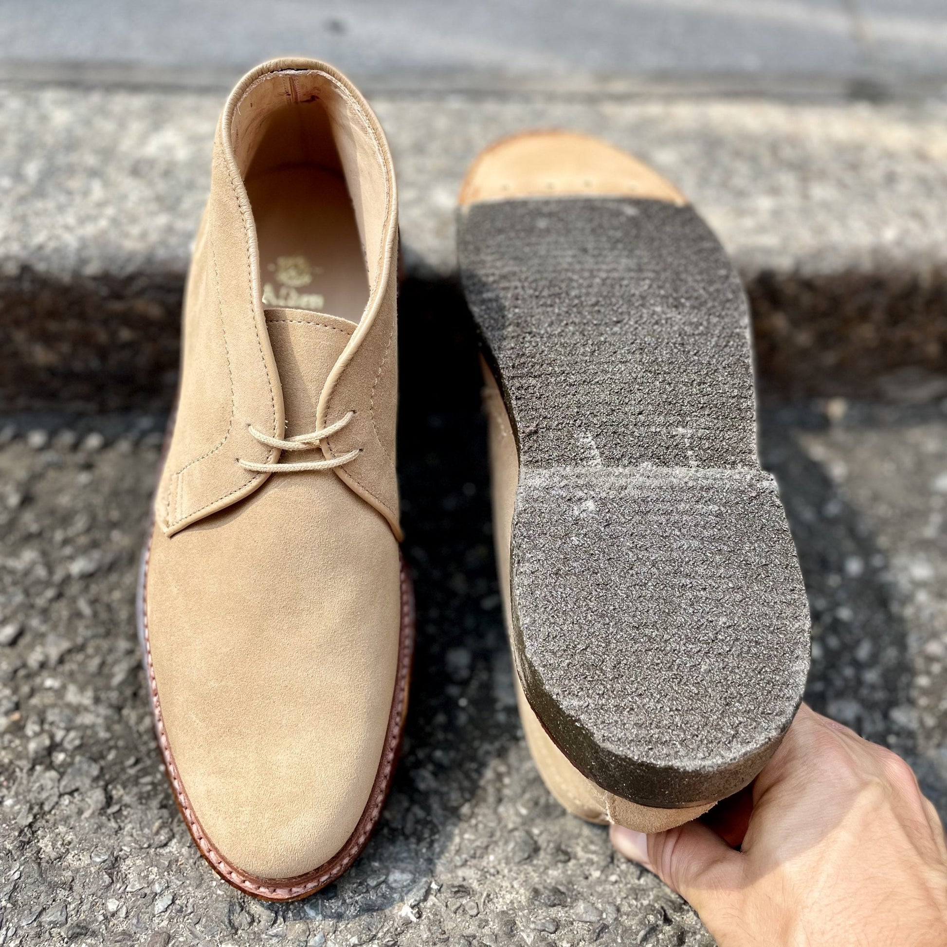 A pair of Alden Madisons 14580 - Desert Chukka Boot in Tan Suede is placed on a concrete surface. One boot stands displaying its profile and laces, while the other lies sole up, showcasing its unique crepe sole texture as a hand securely holds the inverted boot.