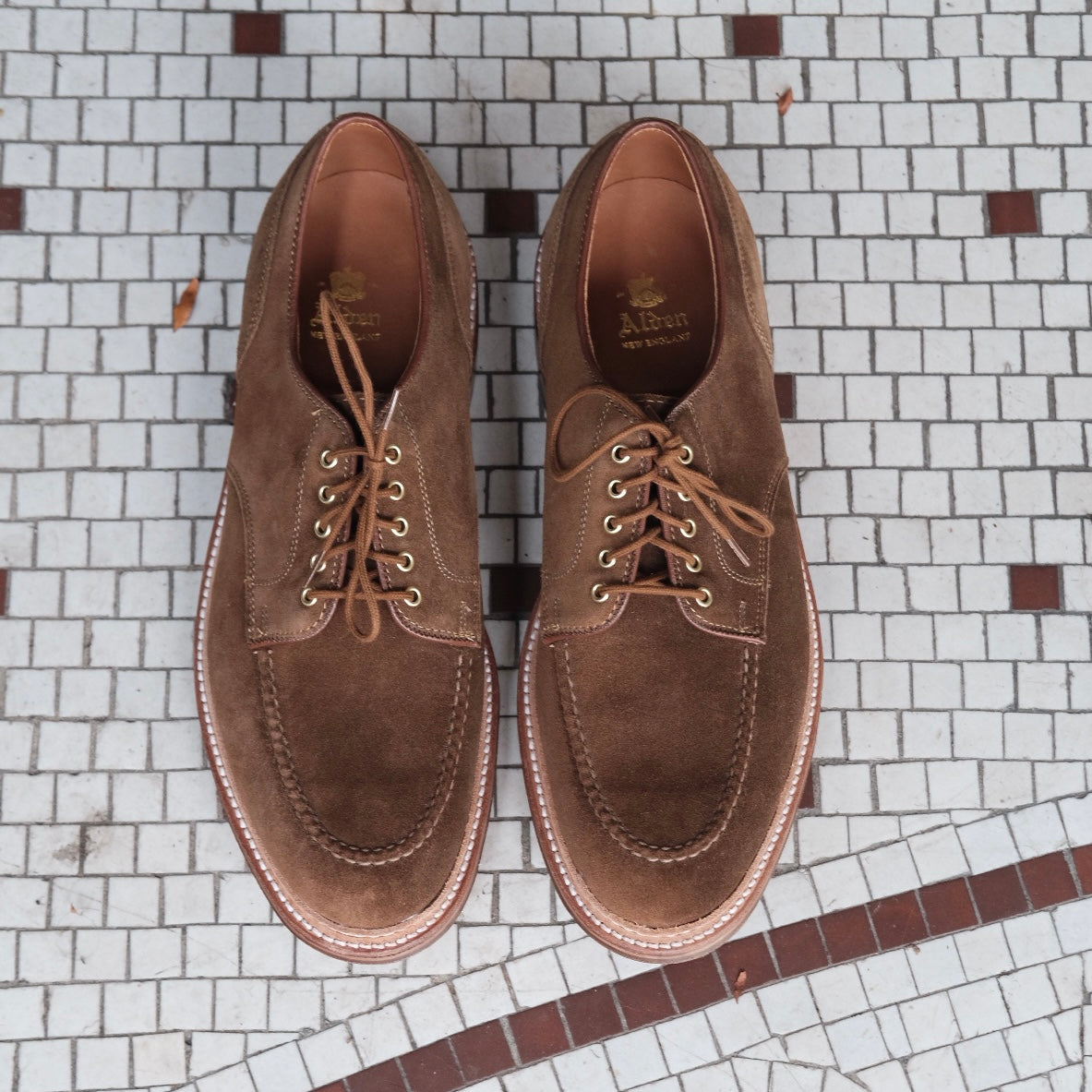 A pair of Alden Madisons 24082 - The Quickley in Snuff Suede shoes with leather lining sits on a tiled floor with a gray geometric pattern and curved brown line.