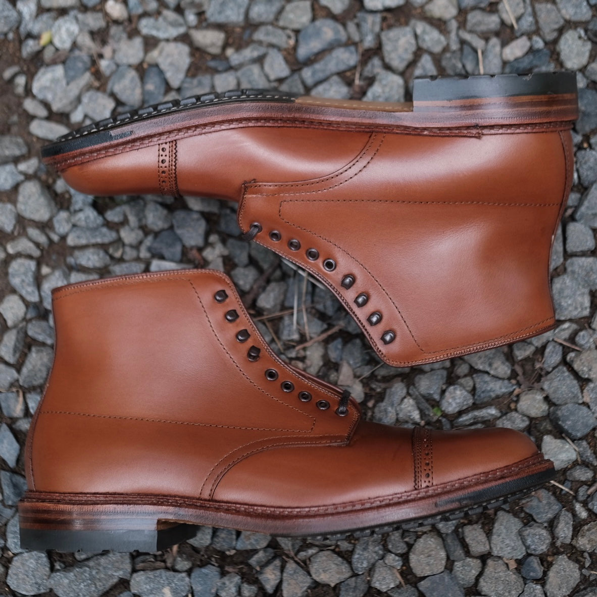 A pair of Alden Madisons D2825HC Jumper Boots in burnished tan calfskin with black eyelets and commando soles rests on a gravel surface, highlighting the craftsmanship and design of the classic Barrie Last with exquisite stitching details from a side view.