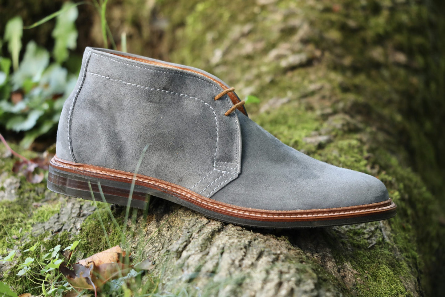 The Alden Madison 1592L Chukka Boot with L3 Sole in Lead Suede, featuring brown laces and stitching, rests on a moss-covered log outdoors amid lush greenery.
