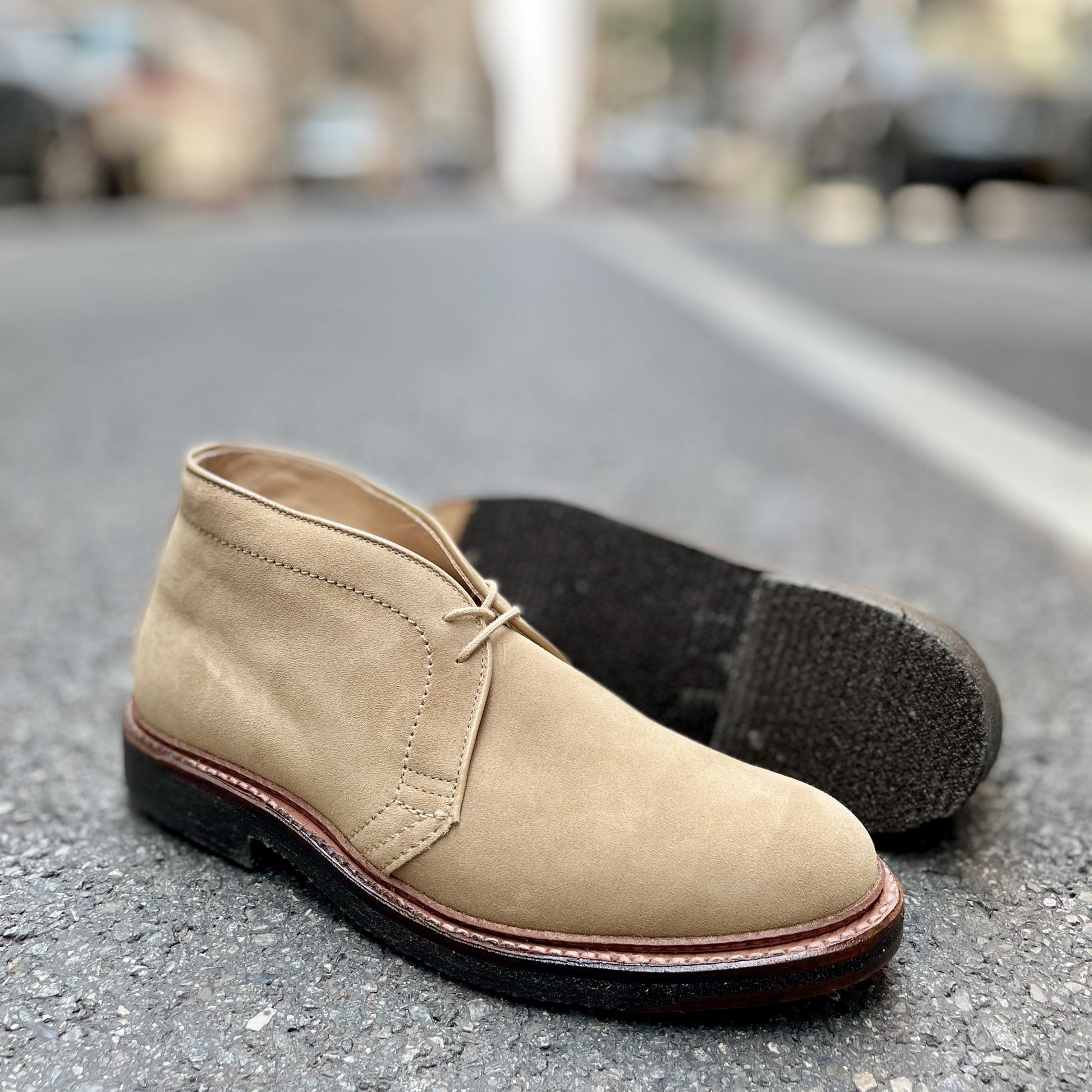 A pair of Alden Madison Desert Chukka Boots (model 14580) in tan suede is on an asphalt road. One boot stands, while the other lies on its side, revealing the unique dark crepe sole and textured heel against a blurred street backdrop.