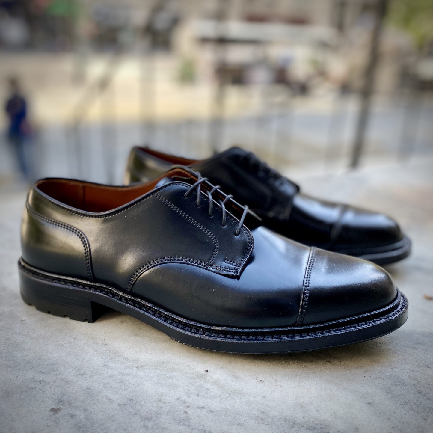 A pair of Alden Madisons 2175C - DeRosa Captoe Blucher shoes in Black Shell Cordovan are placed on a concrete surface with a blurred outdoor background. These elegant black leather dress shoes feature black laces and a classic design, ideal for any formal event.