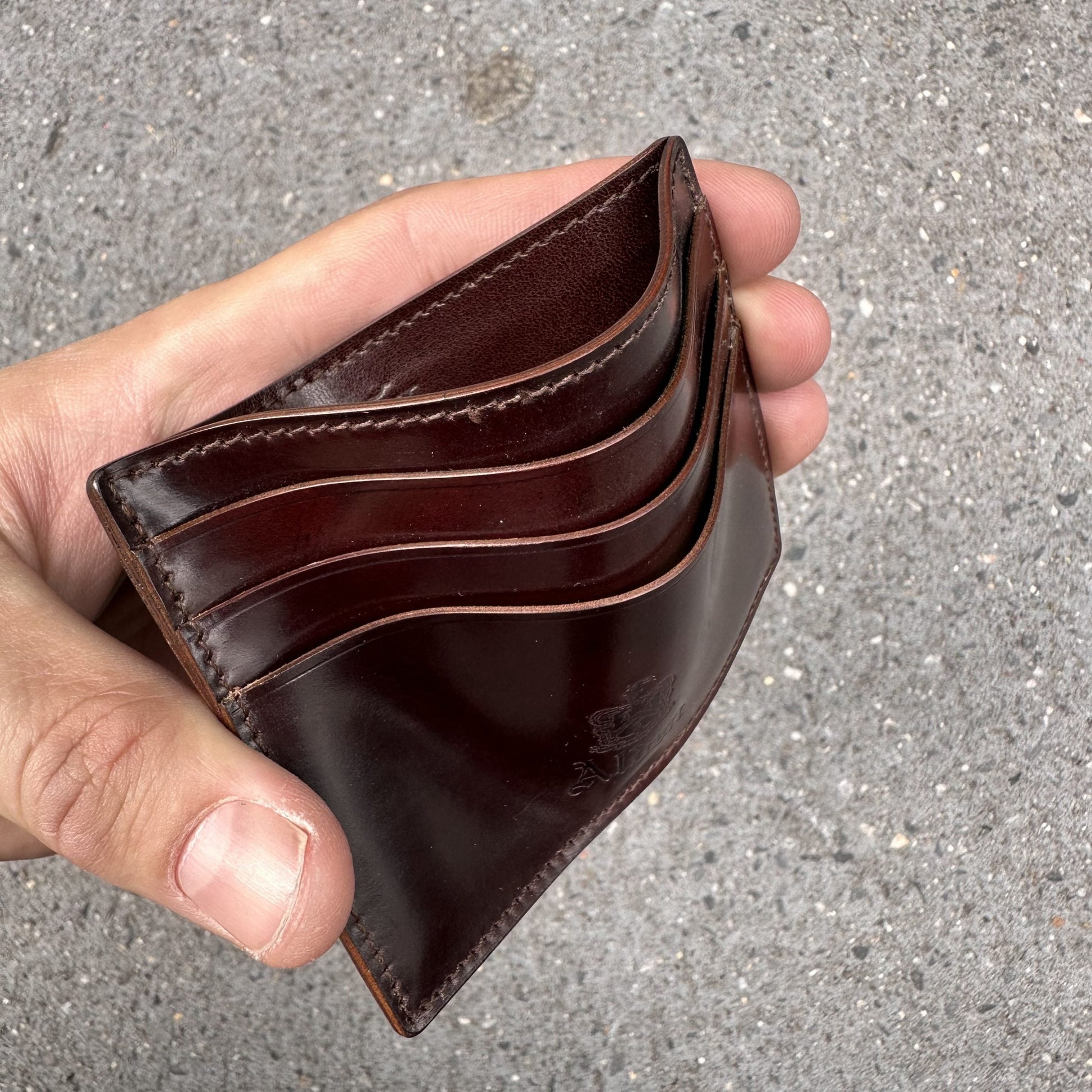 A hand holds an LG 6008 - Alden Color 8 Shell Credit Card Holder by Alden Madison, a brown leather cardholder with three curved slots. It rests on a textured gray surface, highlighting its elegant design.