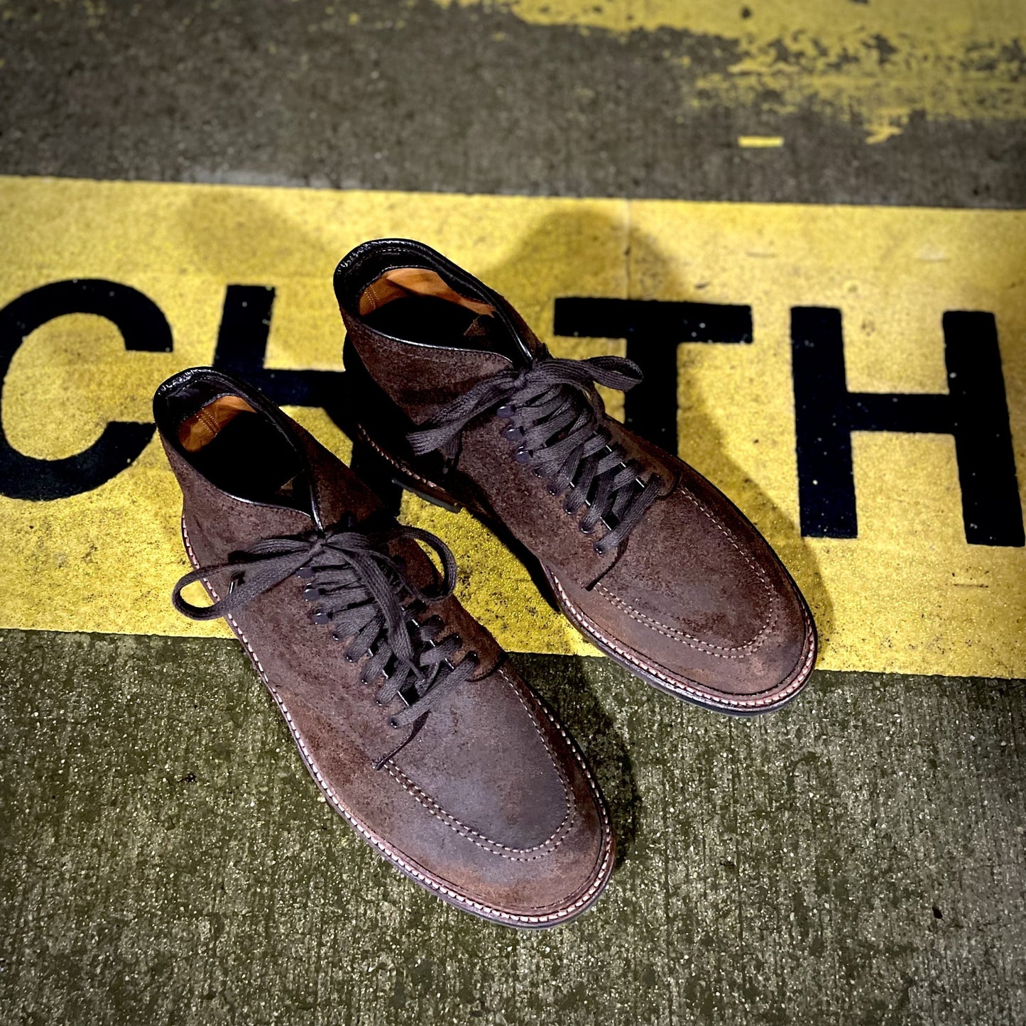 A pair of Alden Madisons 4015HC - Indy Boot in Tobacco Reverse Chamois, distinguished by dark laces and a rugged Commando Sole, sit on concrete partially covering black letters on a yellow background—ideal for both city streets and rustic trails.