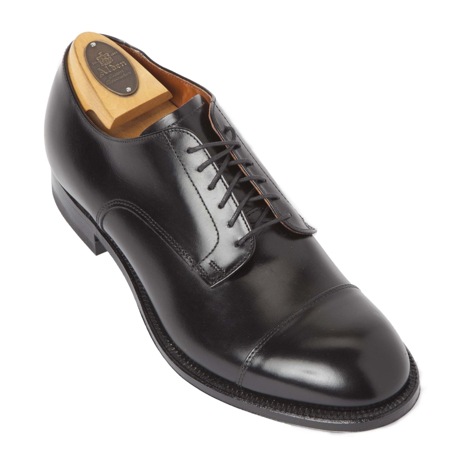 The Alden Madison 558 is a sleek black calfskin dress shoe with a modified cap toe and Single Oak Leather Sole. Black laces enhance its elegance, while a wooden shoe tree maintains its shape, all set against a plain white background.