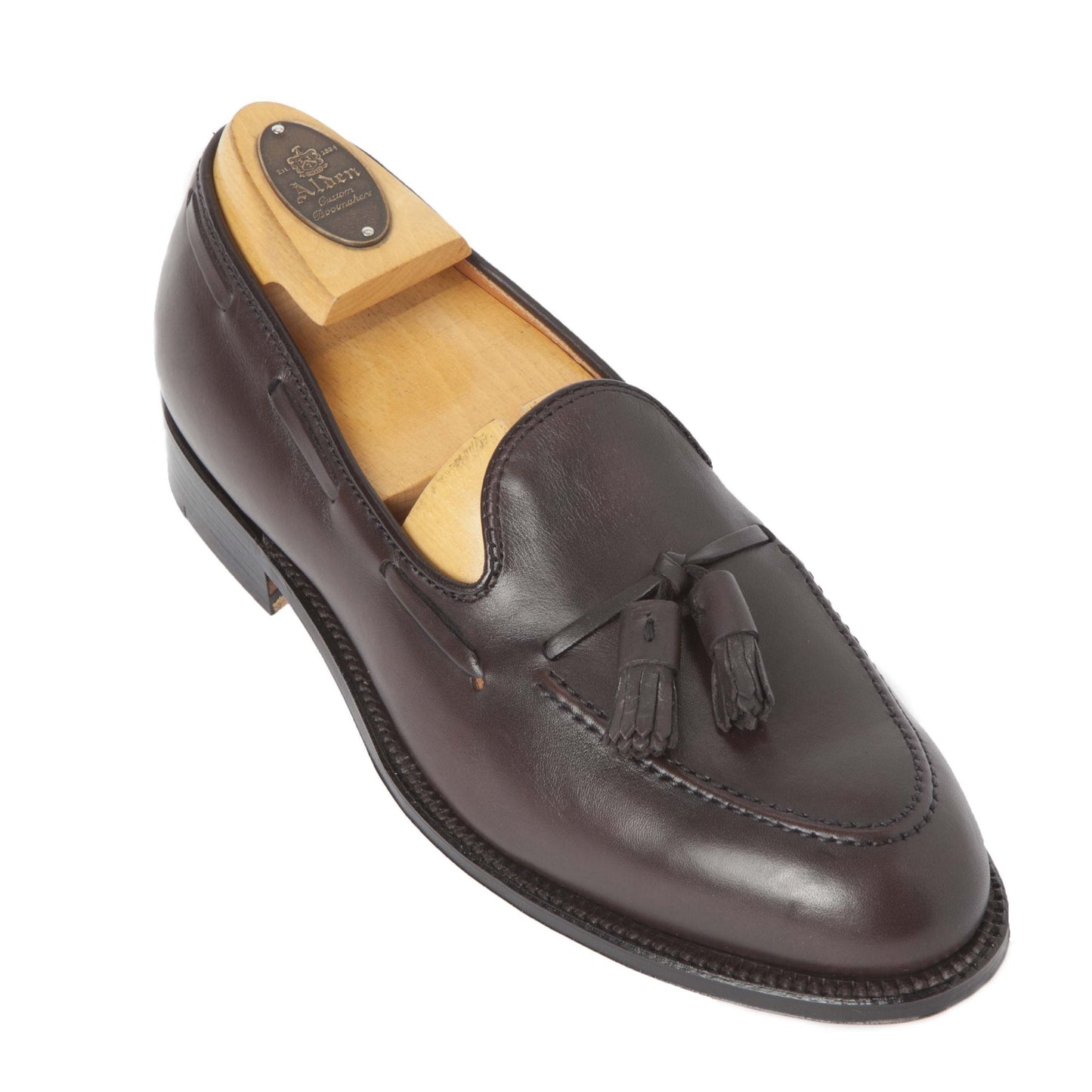 The Alden Madison Tassel Loafer (Model 561), made from dark brown calfskin, showcases detailed stitching, two tassels, and a wooden shoe form inside. Crafted on an Aberdeen Last, it features a light tan interior and a smooth leather sole.