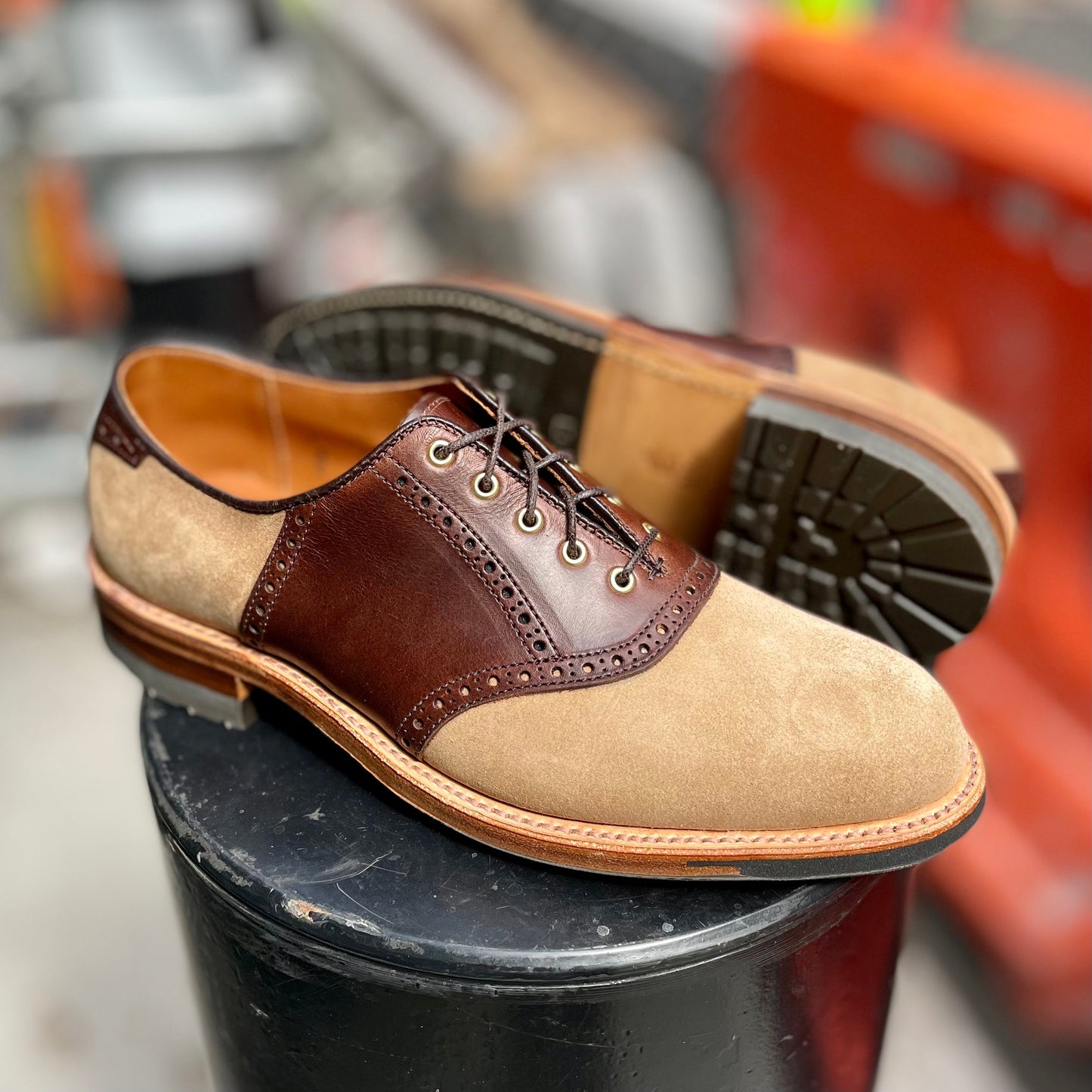 A pair of stylish Alden Madison D1304C - Valhalla Saddle Oxford shoes in brown CXL leather and tan suede rest on a black circular surface, with one upright and the other leaning behind, enhancing their elegance against a blurred background.