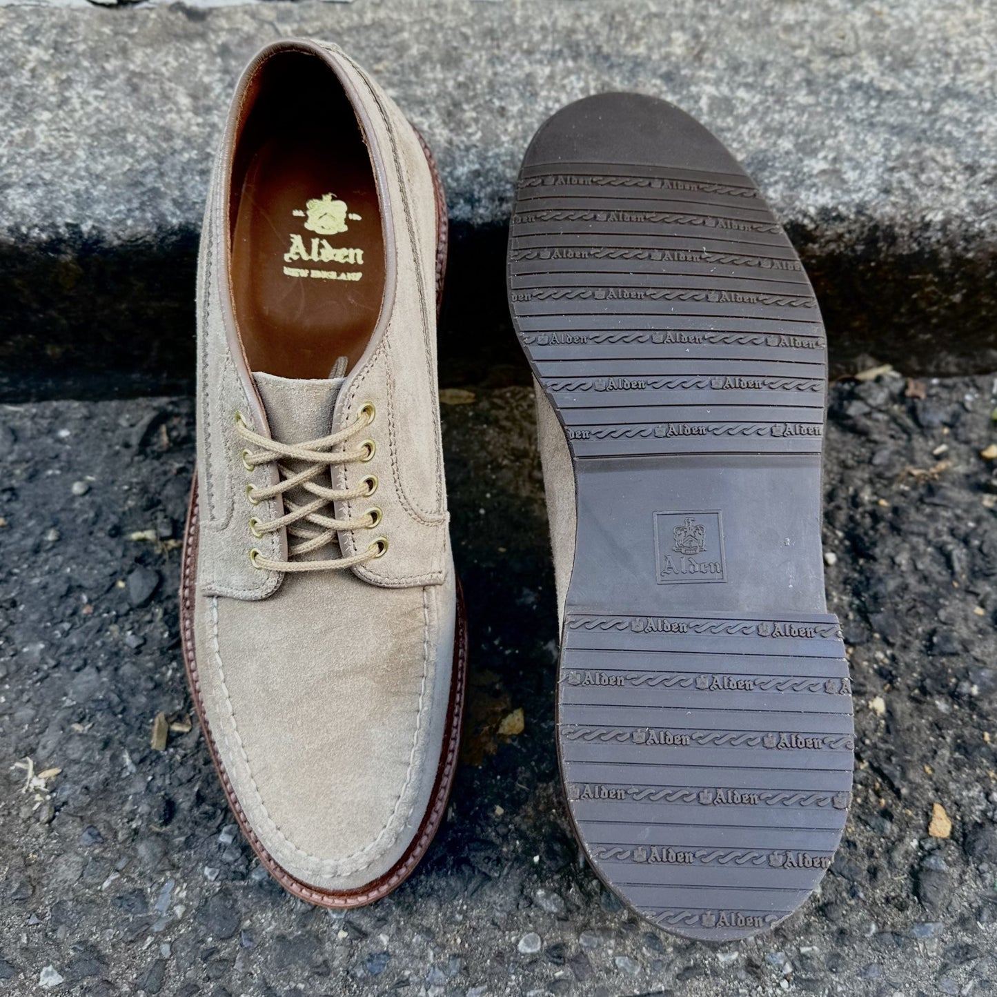 A pair of Alden Madison D1634L - Ranger Mocc in Milkshake Suede on a rugged outdoor surface: one upright, showing laces and logo inside, the other flipped to reveal its Brown Rubber Sole with brand markings.