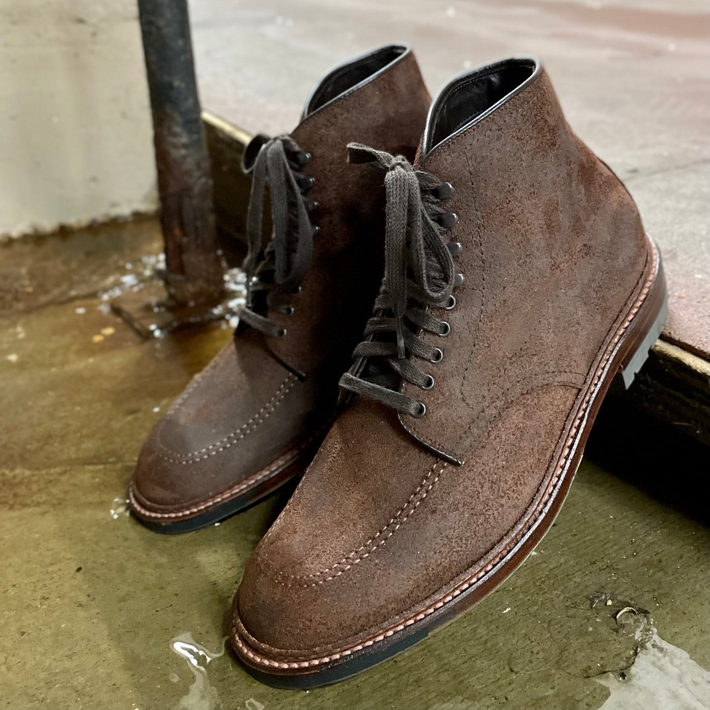 On a wet concrete surface, the Alden Madison 4015HC - Indy Boot in Tobacco Reverse Chamois stands with black laces and Commando Soles. Industrial elements like a puddle and metal pipe create the backdrop.