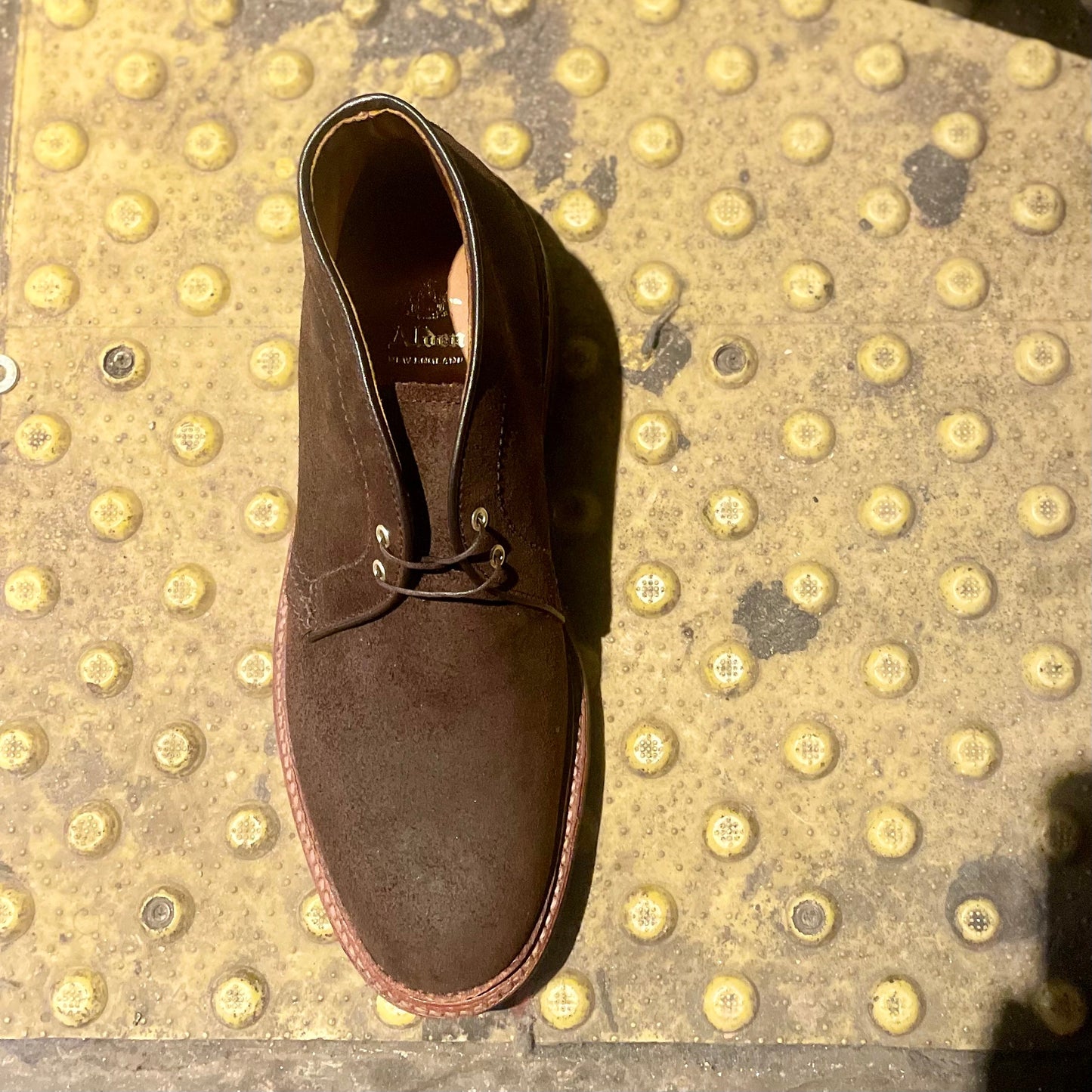 The Alden Madison D1717C Catskills Chukka Boot in Tobacco Reverse Chamois, featuring a Commando sole, is displayed on a yellow textured surface with raised bumps typically found at train platform edges for tactile warning.
