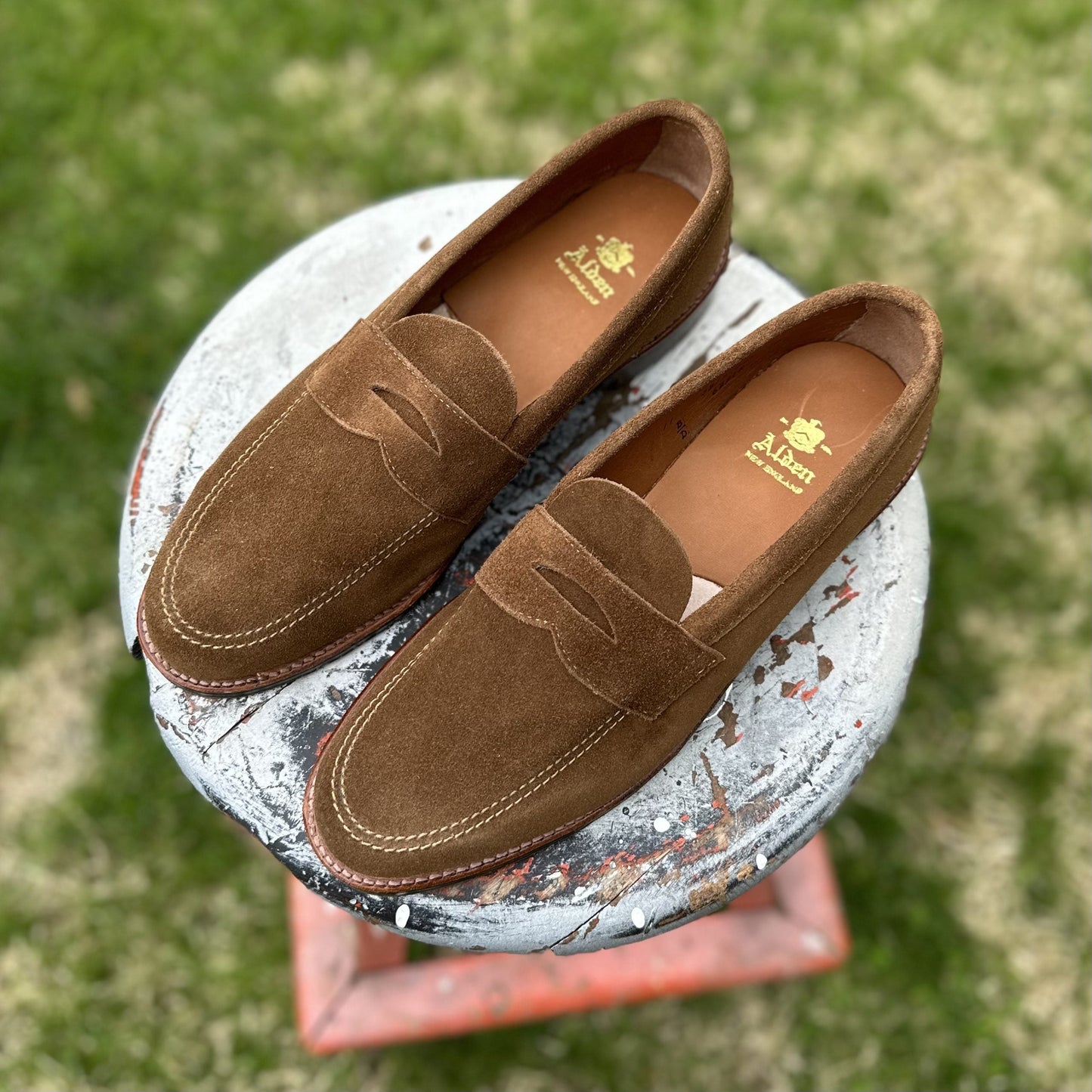 The Alden Madison 6221L - Unlined Penny Mocc in Snuff Suede, featuring light stitching and a mocc toe, rests on a weathered round stool against a grassy outdoor backdrop.