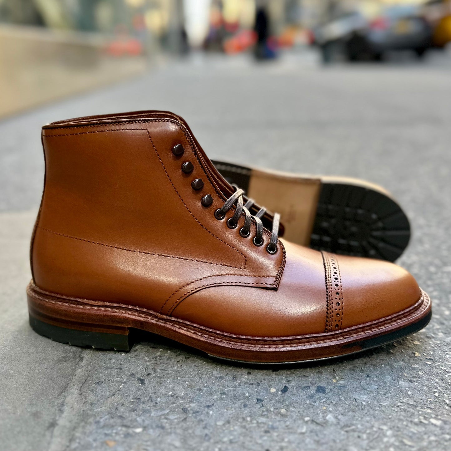 A pair of Alden Madisons D2825HC Jumper Boots in burnished tan calfskin, featuring a cap toe design and commando sole with black laces, sits on concrete. One boot leans against the other, set against a blurred urban backdrop.