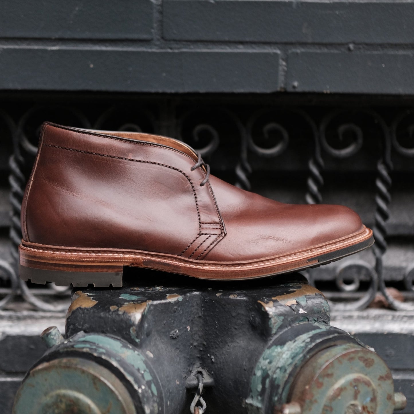 The Alden Madison 13781C Chukka Boot in Brown Chromexcel, featuring a Commando sole, is showcased on a weathered metal surface with a dark backdrop of wrought iron accents and green painted bricks.