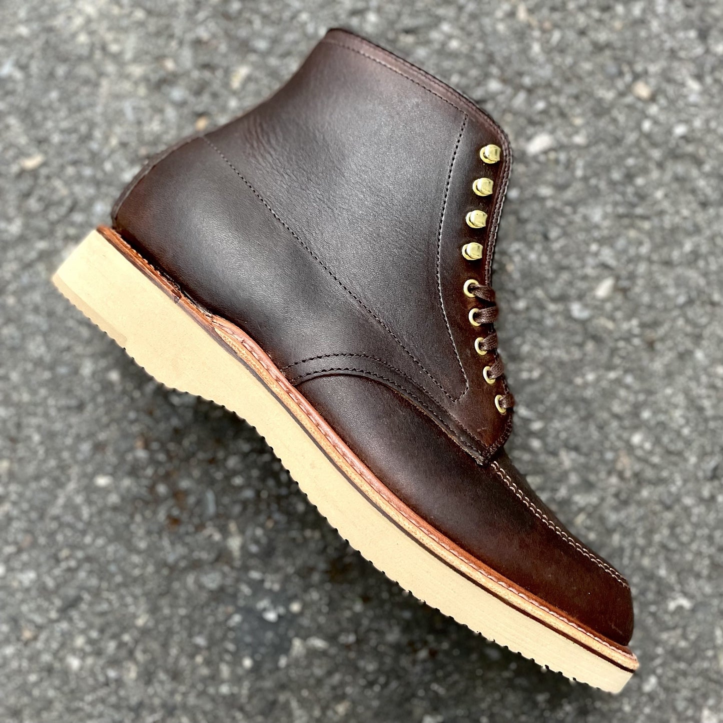 The Alden Madison D1926H Porter Indy Boot, crafted from antique tan Kudu leather with gold eyelets and a tan sole, rests stylishly on the gray asphalt. Its sleek design and craftsmanship make it ideal for any adventure.