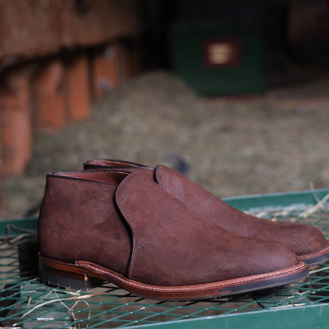 The Alden Madison D8704C - Folsom Slip On Chukka in Tobacco Reverse Chamois, featuring Commando rubber soles, is displayed on a green metal surface with rustic barn and hay accents in the background.