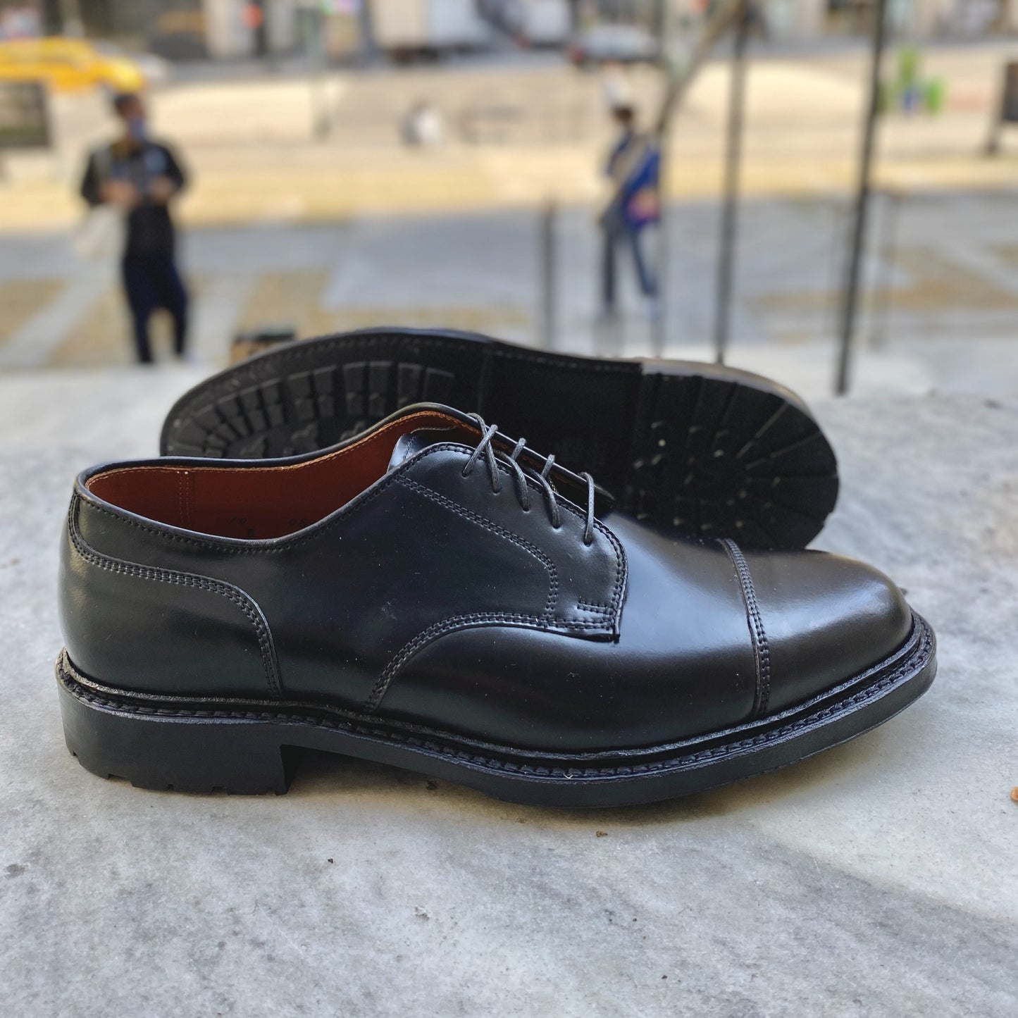 The 2175C - DeRosa Captoe Blucher in Black Shell by Alden Madison is showcased on a stone surface. The blurred background features people and an urban sidewalk scene, with the shoes Commando Sole visible, highlighting its tread pattern.