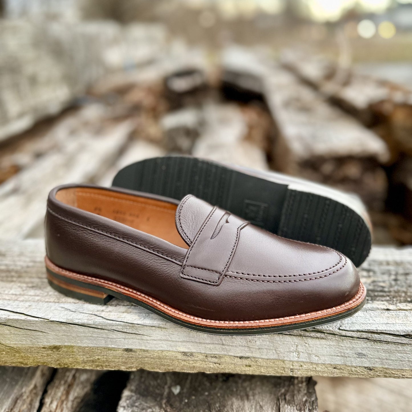 The D2203L - Penny Loafer in Brown Lady Calf by Alden Madison rests elegantly on a wooden surface. Its black rubber sole is visible, highlighting the side profile and bottom design, while the blurred background enhances the rustic wood texture.