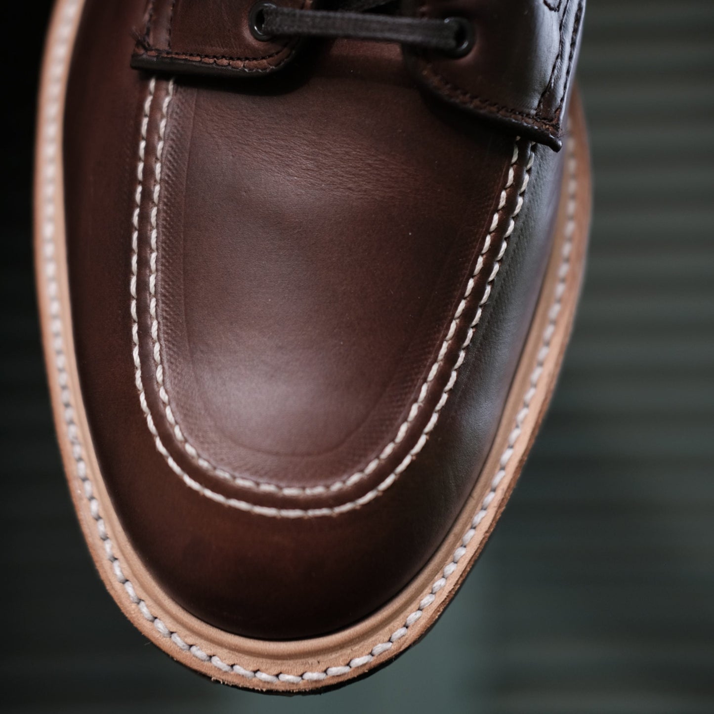 Close-up of the Alden Madison 403C - Indy Boot w Commando Sole in CXL, highlighting its detailed stitching around the toe on rich brown Chromexcel leather. The polished texture and light tan sole edge crafted on a Truebalance Last stand out against a blurred horizontal line background.