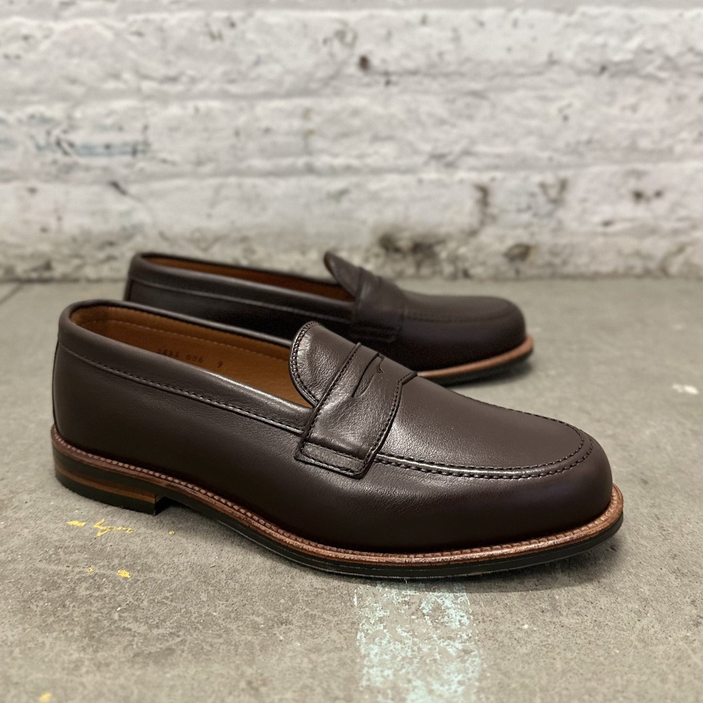 A pair of Alden Madisons D2203L - Penny Loafer in Brown Lady Calf, featuring an antique flat welt and stitching detail, rests on a gray concrete surface against a textured white brick wall, highlighting their classic design.