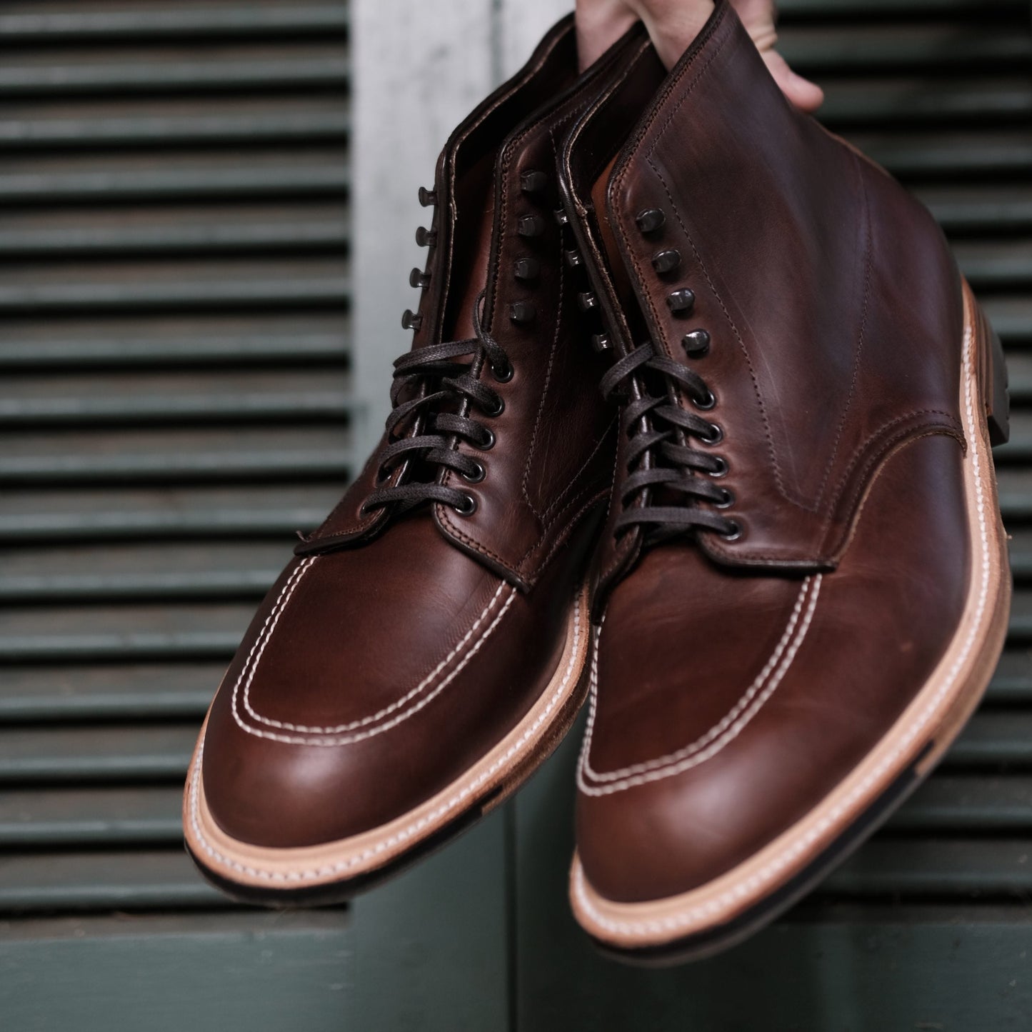 A hand holds a pair of Alden Madisons 403C - Indy Boot in Chromexcel leather, featuring white stitching, the Truebalance Last, and a rugged Commando Sole against a green slatted surface.