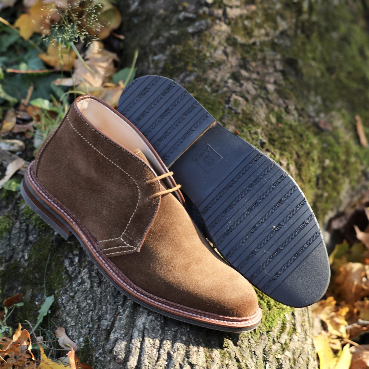 The Alden Madison 1593L Chukka Boot with L3 Sole in snuff suede sits on a tree stump surrounded by fallen leaves. One boot is upright, and the other showcases its rubber soles patterned grip. Sunlight enhances the textured suede.