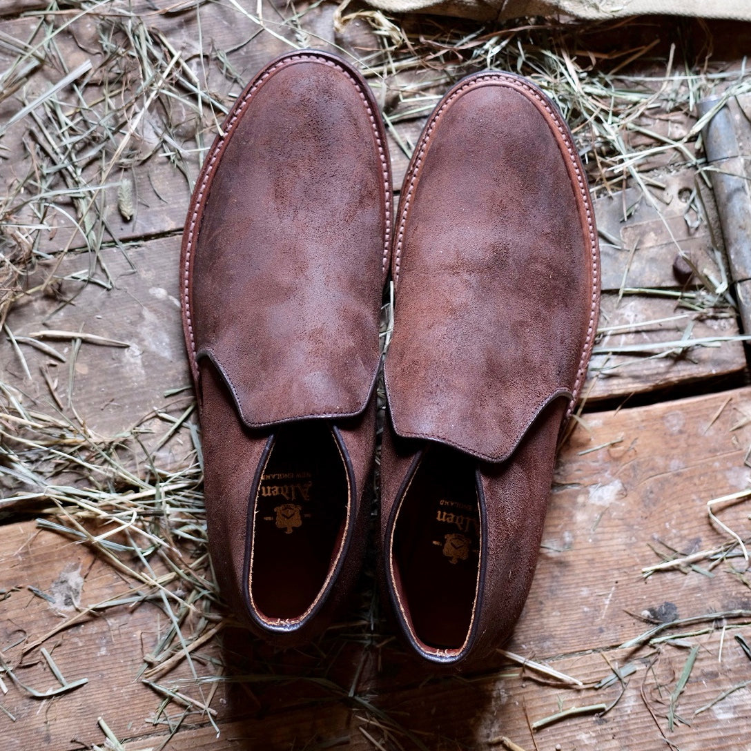 The Alden Madison D8704C - Folsom Slip On Chukka in Tobacco Reverse Chamois, featuring a sturdy commando rubber sole, sits side by side on a rustic wooden floor strewn with dry hay.