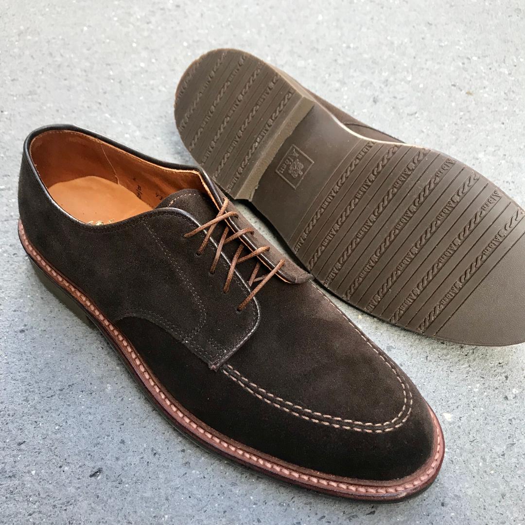 The Alden Madison 704 Mocc Toe Blucher in Brown Suede, featuring light brown dress rubber soles and visible stitching, is shown with one shoe upright and the other on its side to display the textured rubber outsole.