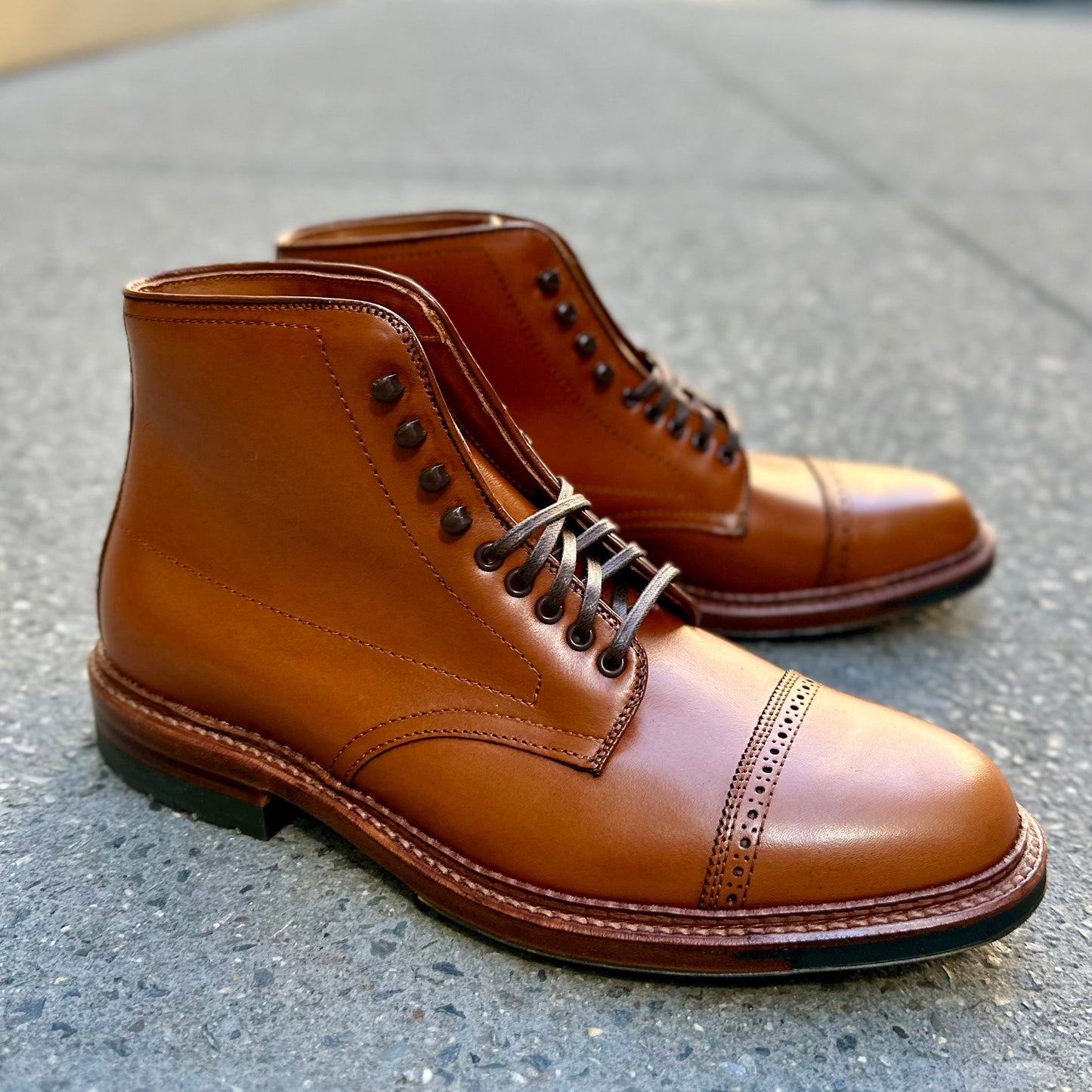 A pair of Alden Madisons D2825HC Jumper Boots, crafted in burnished tan calfskin with cap toes and dark laces, rest stylishly on a textured gray stone. They boast visible stitching, a slight heel, and commando soles for added comfort and modern flair.