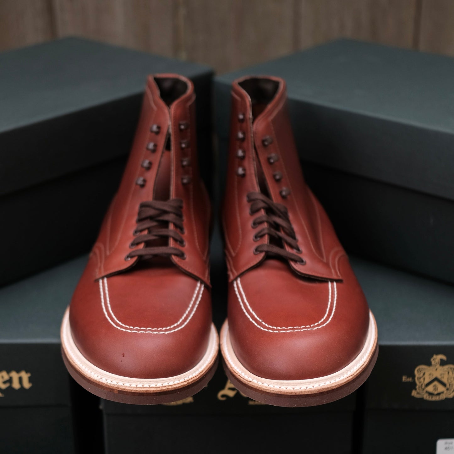 The 405 Indy Boot (Original) in Mid Brown Calfskin by Alden Madison, featuring laces and white stitching with a distinctive contoured design, is crafted from durable leather and displayed atop two green shoeboxes against a wooden background.