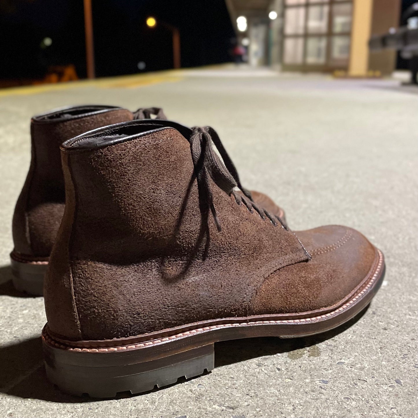The Alden Madison 4015HC - Indy Boot in Tobacco Reverse Chamois, featuring rugged commando soles and visible stitching, sits on a concrete surface as slightly blurred lights and buildings create an urban backdrop.