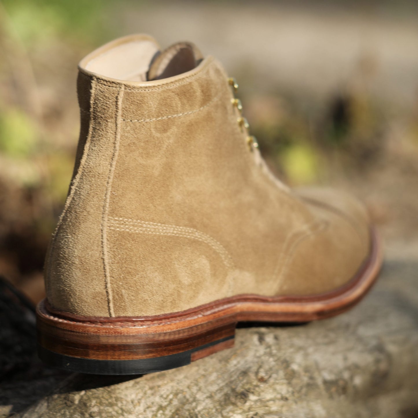 A close-up of Alden Madisons D2685H - 2/2 Cap Toe Boot in tan suede, featuring a wooden sole and metal eyelets, rests on a textured rock surface. The boot is crafted with precision on the Barrie last, with visible stitching and a softly blurred background.