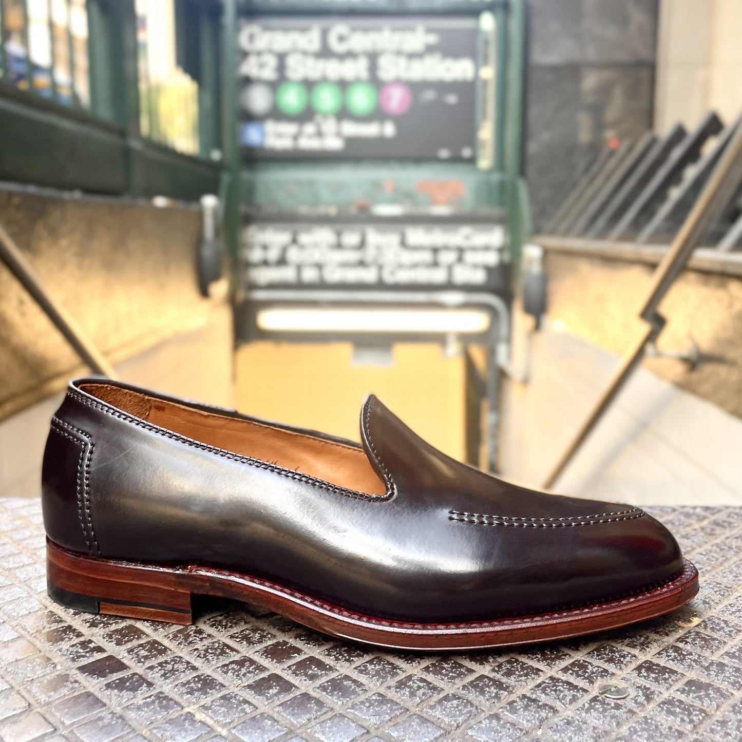 The Alden Madison D1118 - Vanderbilt Slip On in Color 8 Shell, crafted on the Aberdeen Last with a wooden heel, is showcased on tiles near the entrance to Grand Central–42nd Street Station, with subway signage and stairs visible in the background.