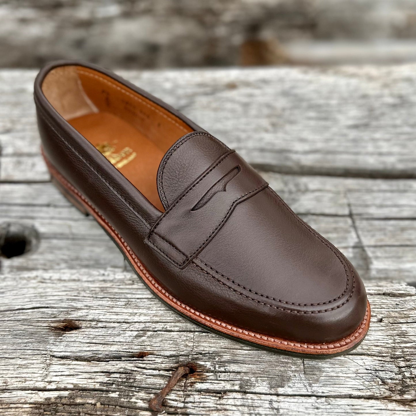 On a textured wooden surface sits an Alden Madison D2203L Penny Loafer in Brown Lady Calf, featuring detailed stitching and a classic penny slot. Its antique flat welt and rubber sole enhance its rustic appeal against the weathered wood background.