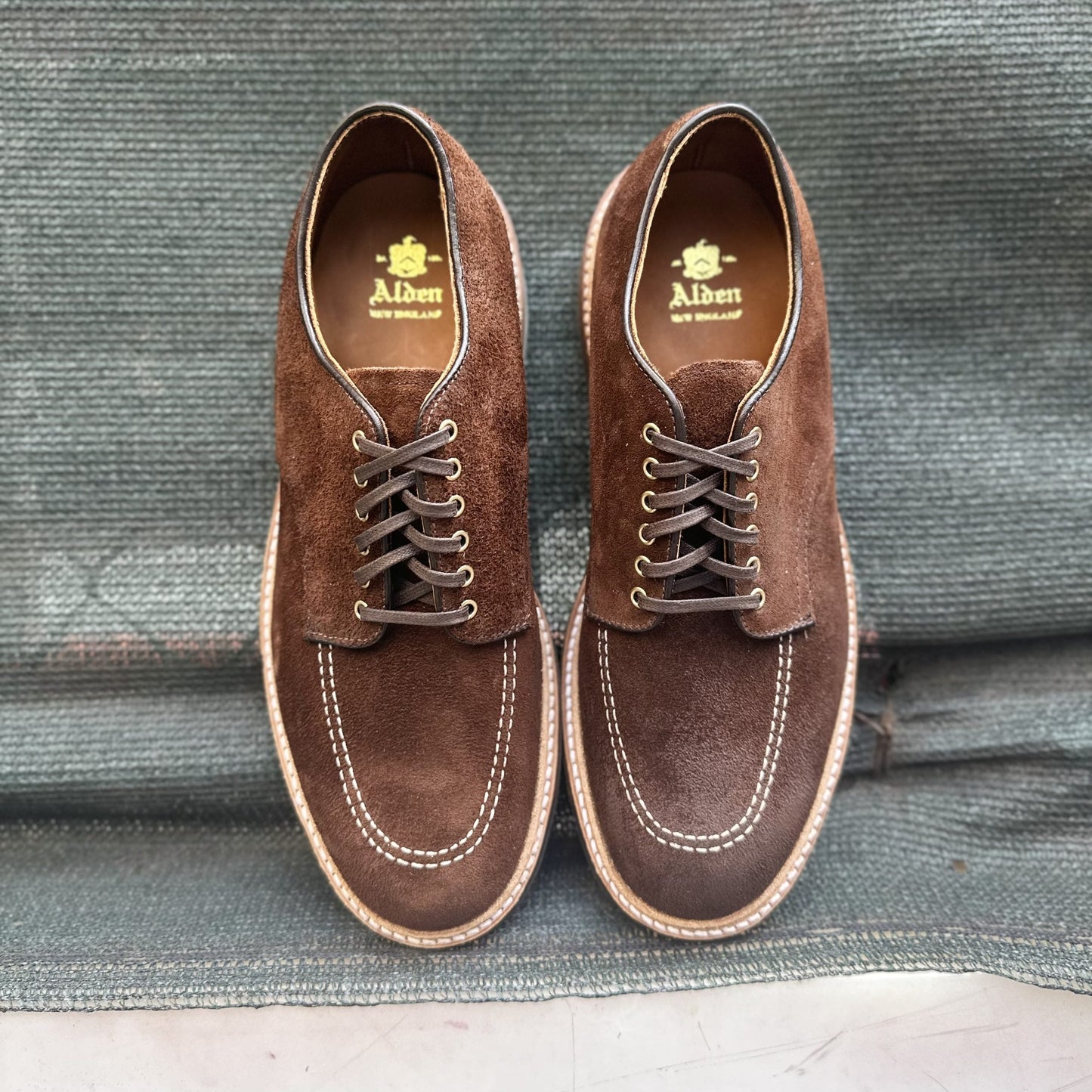 A pair of Alden Madisons D2604C Indy Shoes in Tobacco Reverse Chamois, featuring white stitching and dark brown laces, are placed side by side on a textured gray surface.