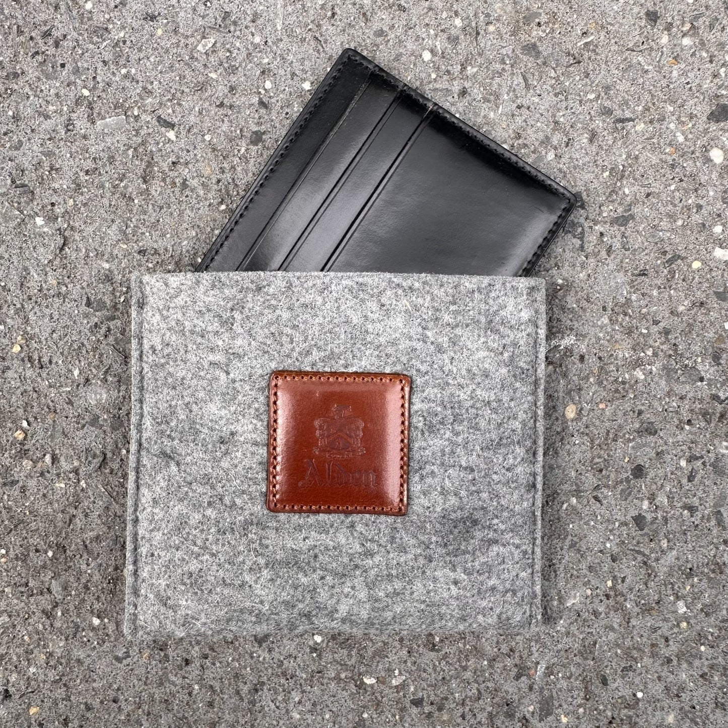 The Alden Madison LG 6009, a sleek black shell cordovan credit card holder, peeks from a gray felt pouch with a brown leather Alden patch. Featuring 6 pockets, it rests against a speckled gray concrete backdrop.