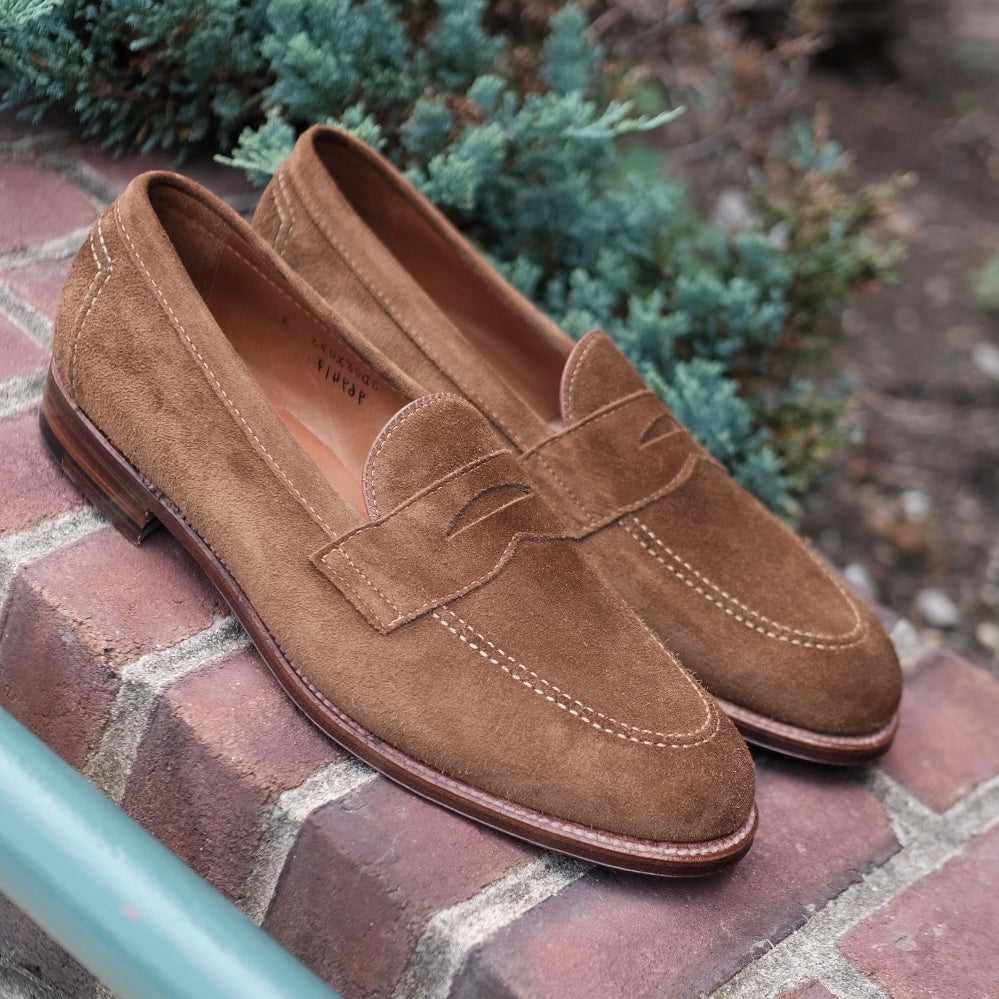 Aldens Madisons 96941F - Flex Penny Loafer in Snuff Suede rests on a brick surface amid green foliage. Built on the Copley Last, these classic loafers feature a polished wooden sole and a small slit detail on the upper.