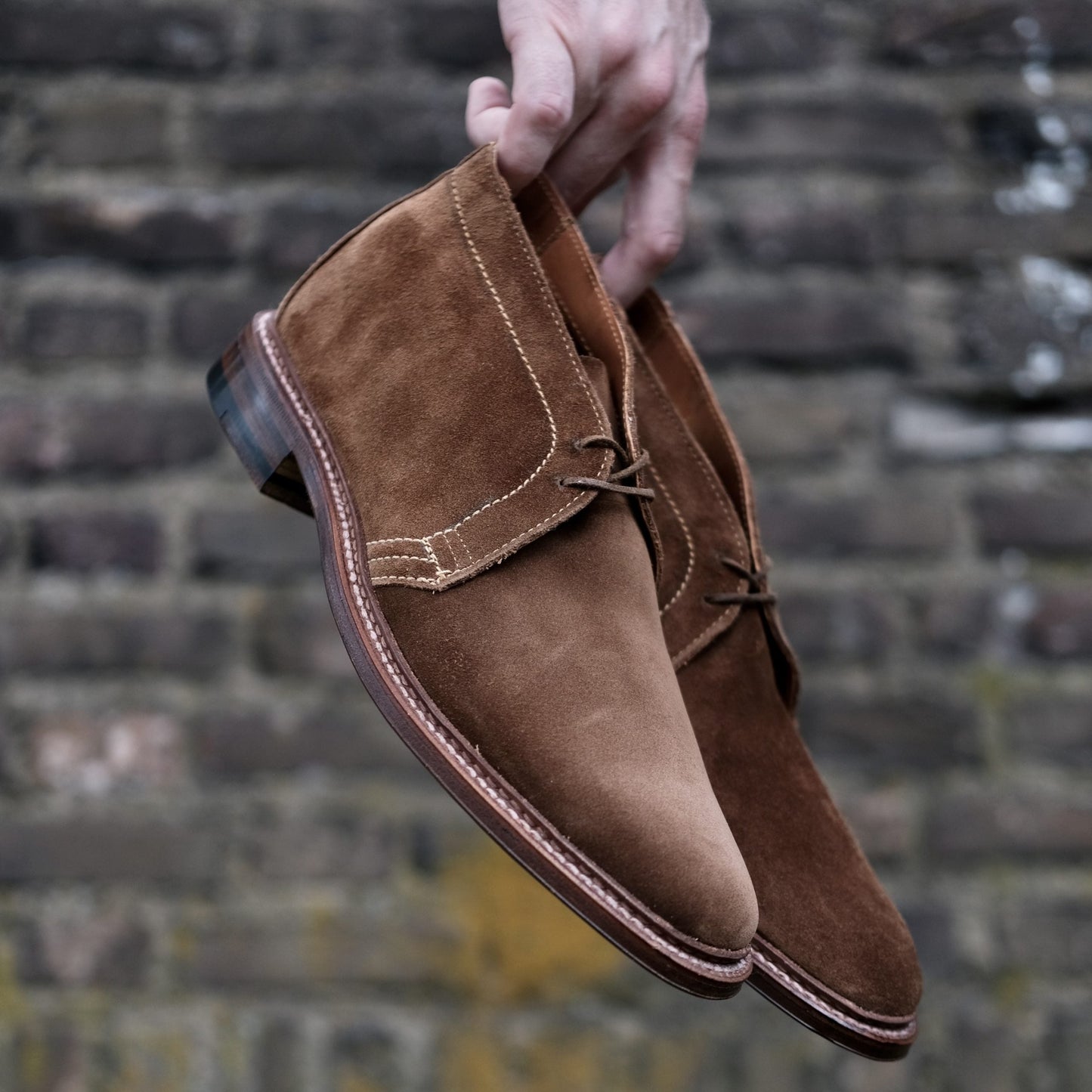 Against a textured stone wall, a pair of Alden Madisons 1493 - Unlined Chukka in Snuff Suede boots crafted on the Leydon Last displays visible stitching and an unlined single flex leather sole with a low heel.