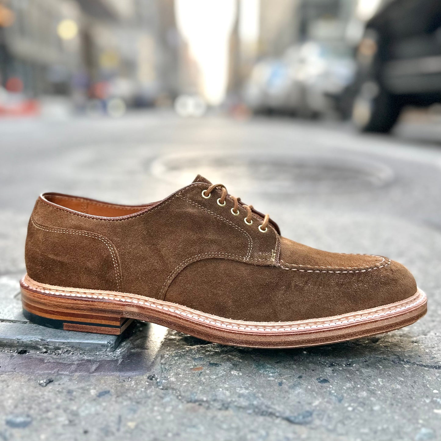 The Quickley in Snuff Suede by Alden Madison, a brown oxford shoe with detailed stitching, is seen on a city street against an urban backdrop, slightly elevated to highlight its timeless elegance.