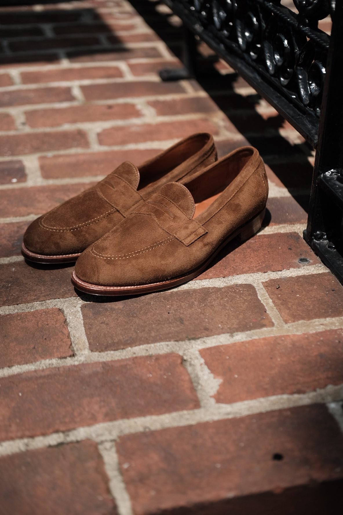 The Alden Madison 96941F Flex Penny Loafer in Snuff Suede rests elegantly on a sunlit brick step, complementing a black wrought iron railing.