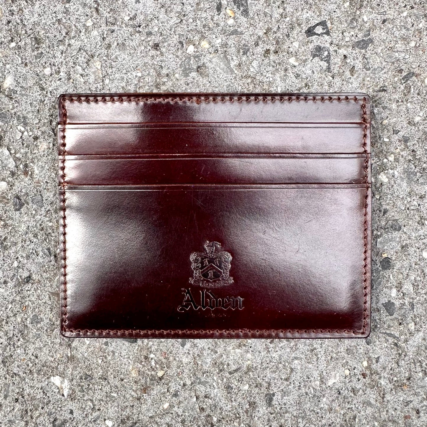 The LG 6008 - Alden Color 8 Shell Credit Card Holder by Alden Madison is a dark brown leather cardholder with three slots, embossed with Albert and a crest. It rests on a textured gray concrete surface, epitomizing refined style and craftsmanship.