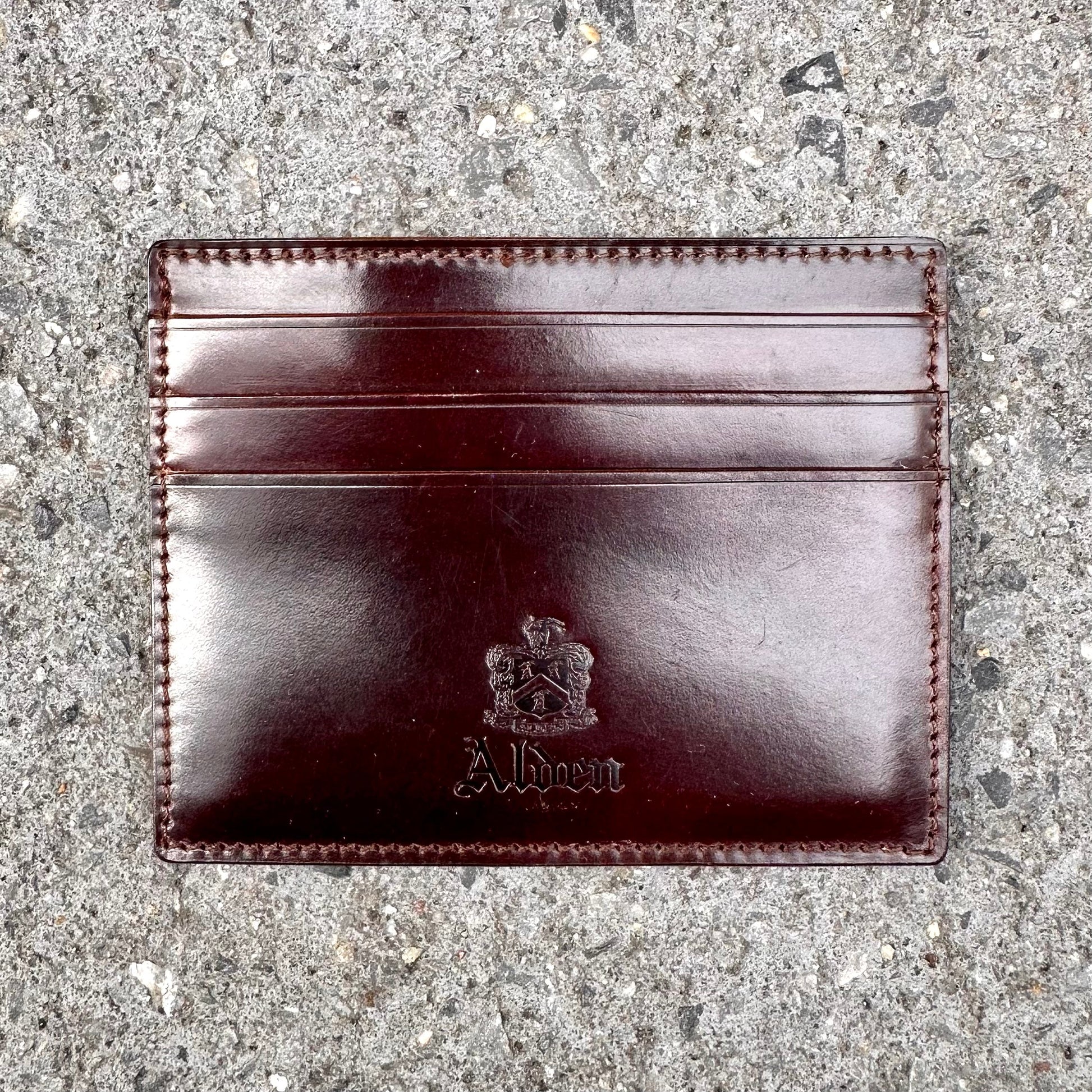 The LG 6008 - Alden Color 8 Shell Credit Card Holder by Alden Madison is a dark brown leather cardholder with three slots, embossed with Albert and a crest. It rests on a textured gray concrete surface, epitomizing refined style and craftsmanship.