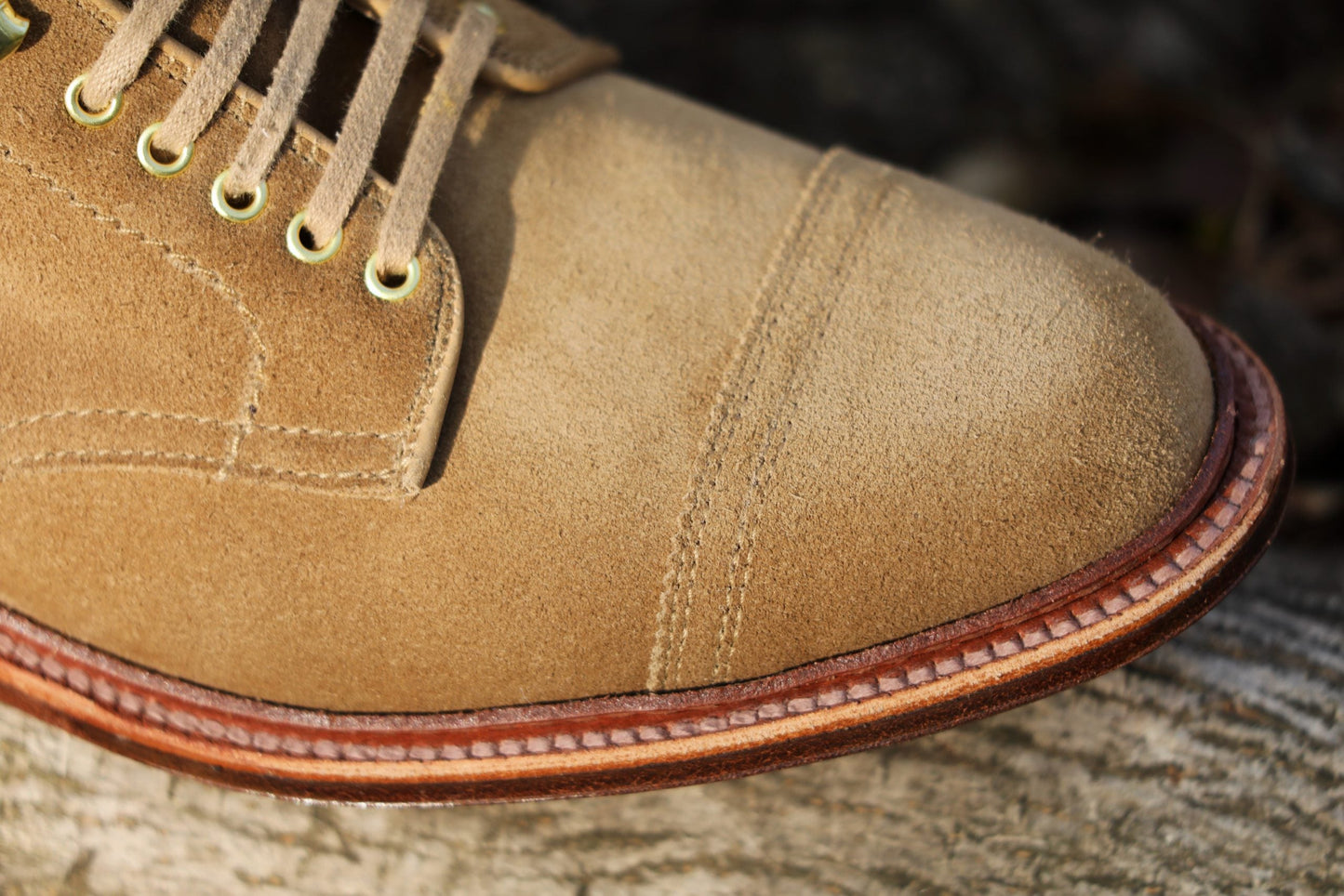 Close-up of the Alden Madison D2685H - 2/2 Cap Toe Boot in Tan Suede with detailed toe cap stitching, brass eyelets, and a welted sole, elegantly resting on a textured surface.